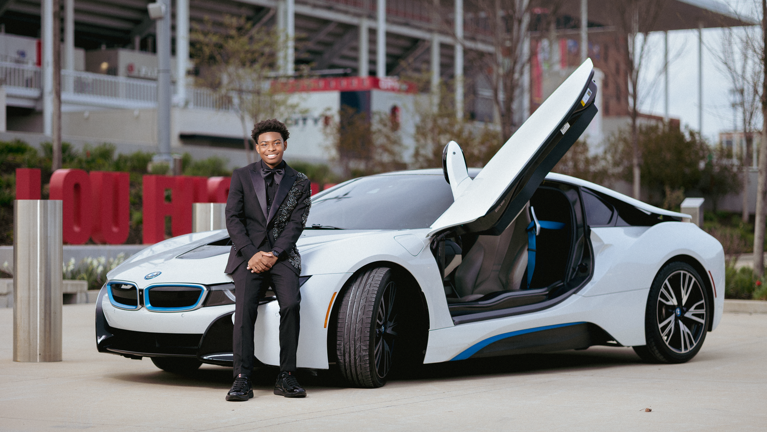 A man in a suit poses beside a white BMW i8 sports car with butterfly doors open in an urban setting.