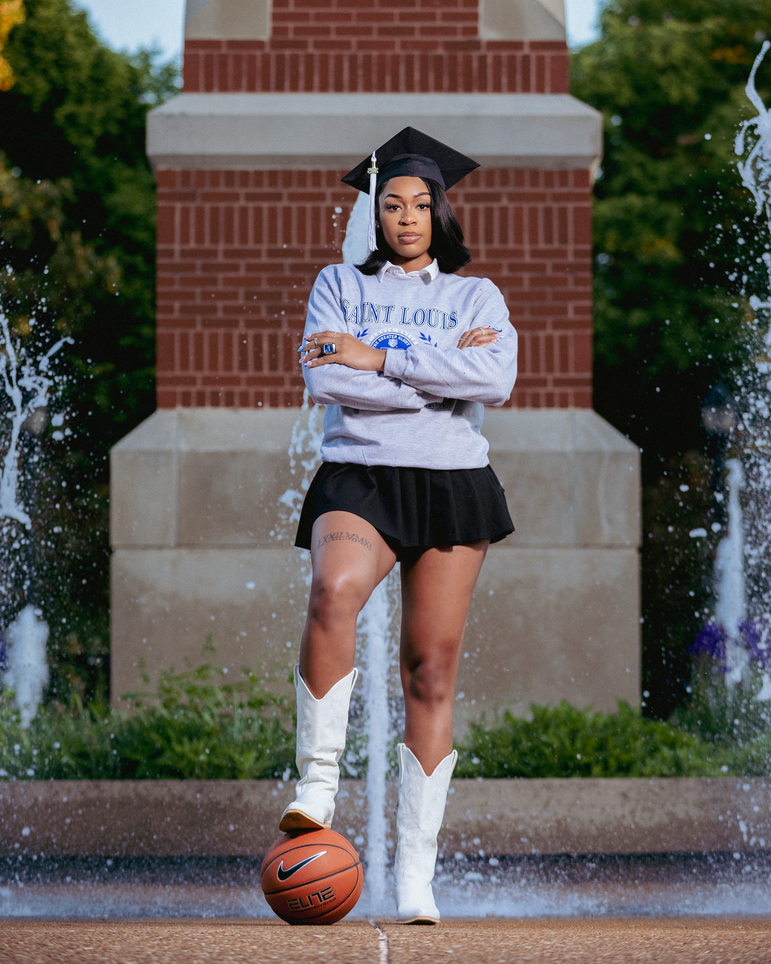 Saint Louis University graduate poses confidently with basketball, wearing cap, sweatshirt, mini skirt, and white cowboy boots by fountain.