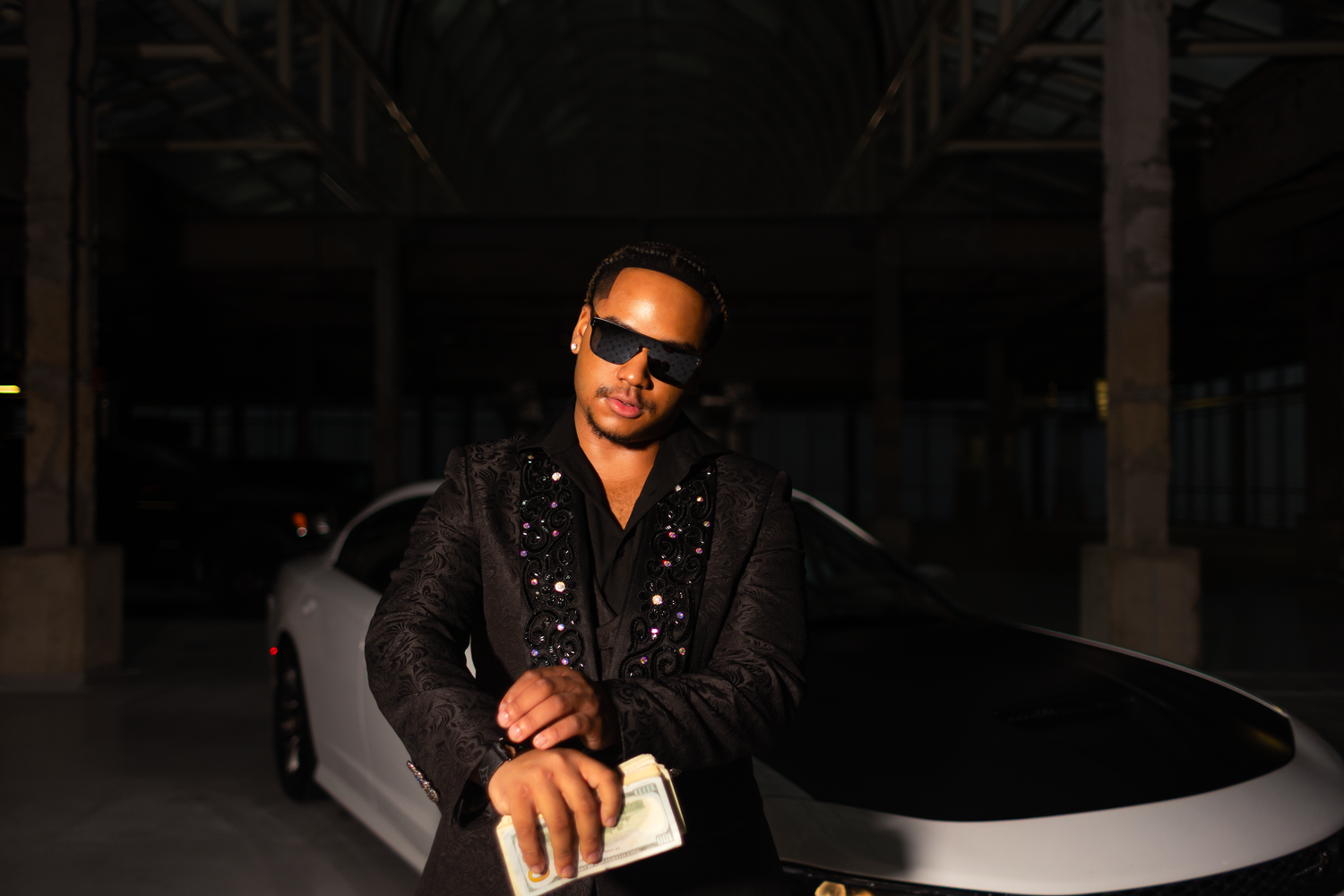 Person wearing sunglasses and black outfit poses near a white vehicle in low light.