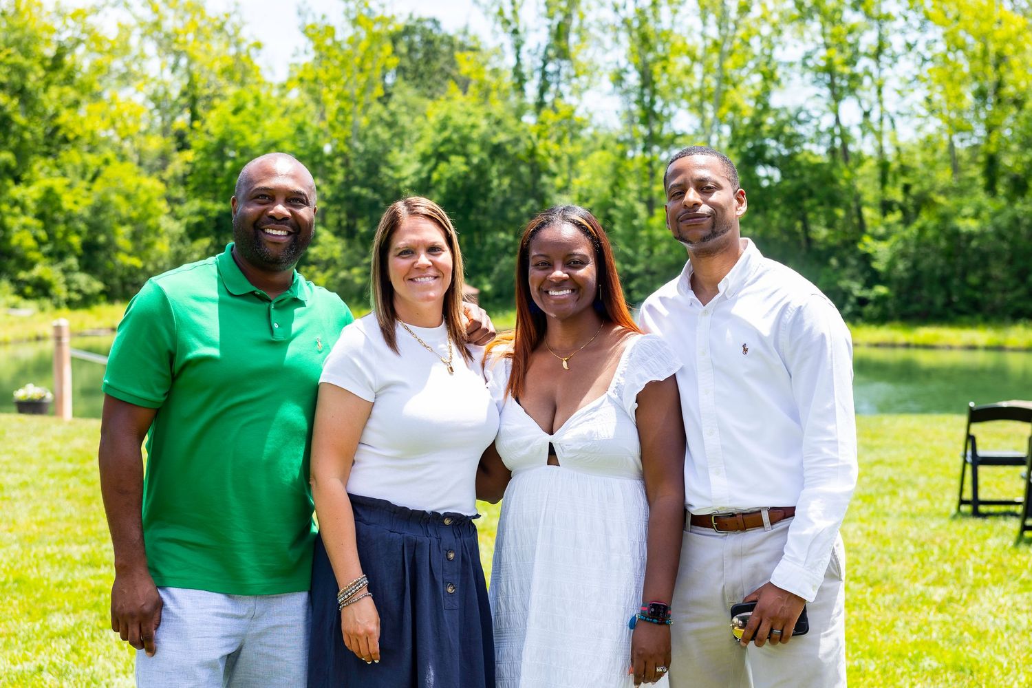 A group of four individuals pose together outdoors on a sunny day with green trees in the background.