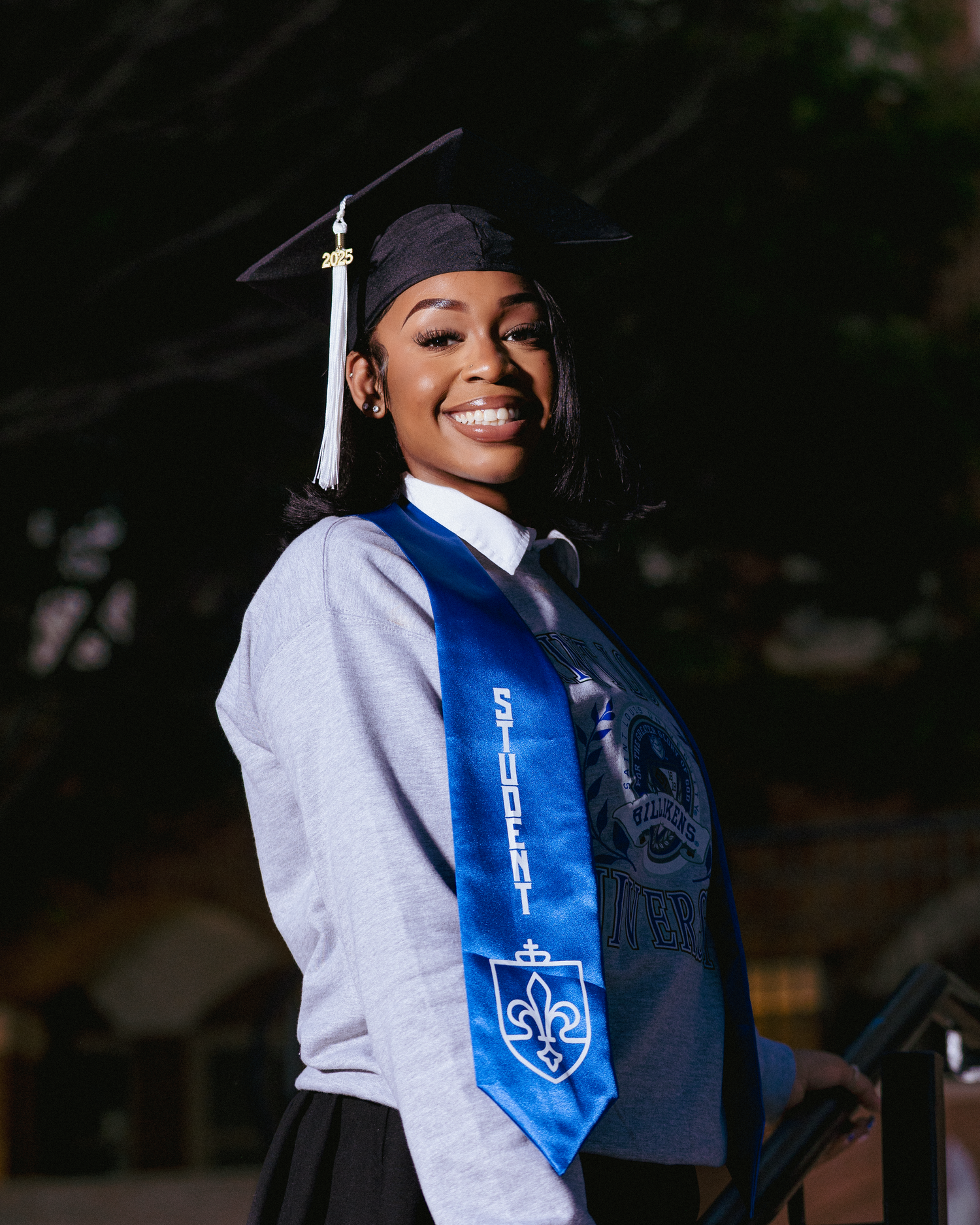 Smiling graduate in cap and gown wearing a blue sash with fleur-de-lis emblem at outdoor ceremony.