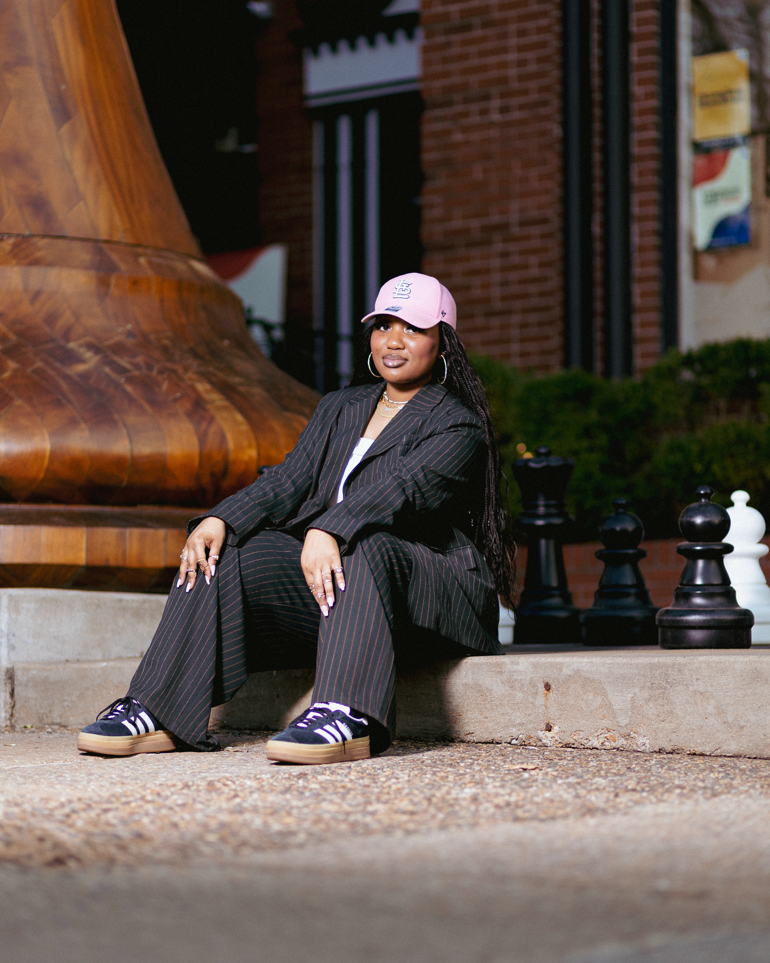 Woman in pinstripe suit and pink cap sits outdoors near large chess pieces and copper fixture.