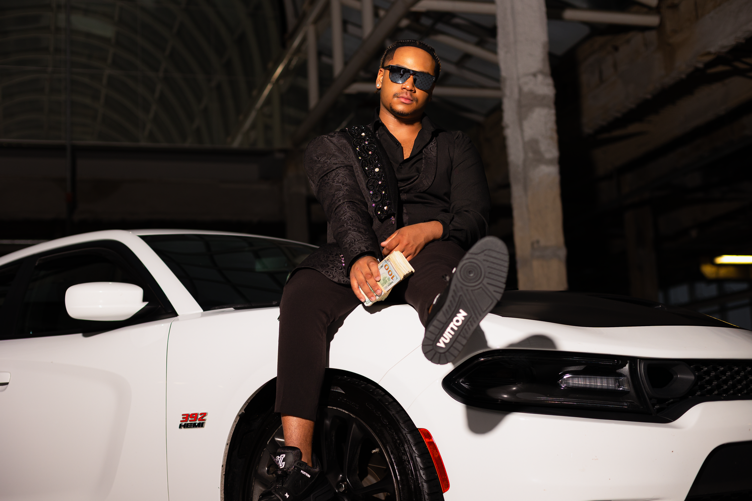 Model in black attire poses dramatically against a white luxury car in a dark parking garage.