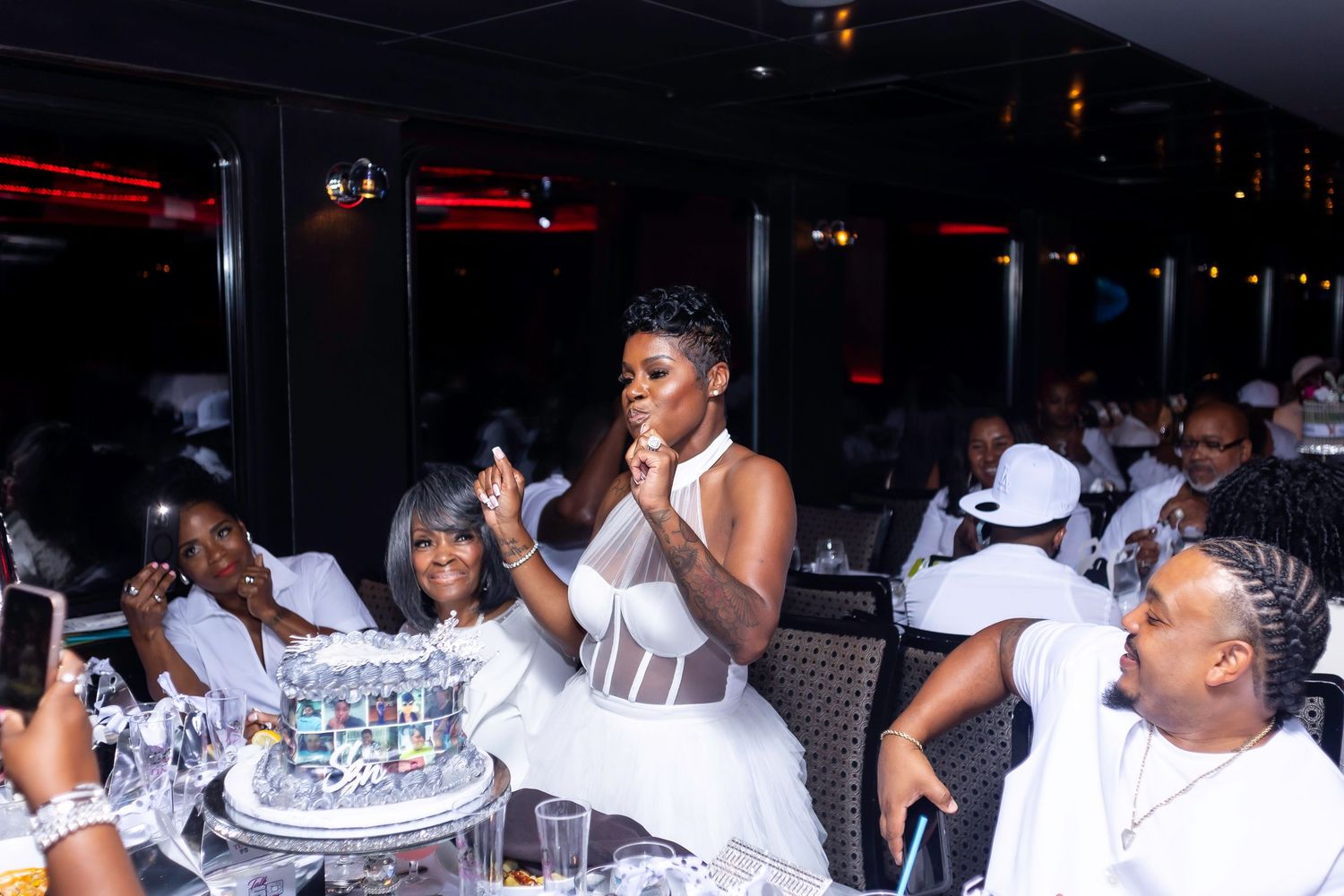 A celebration scene with guests in white attire gathered around a decorated tiered cake.
