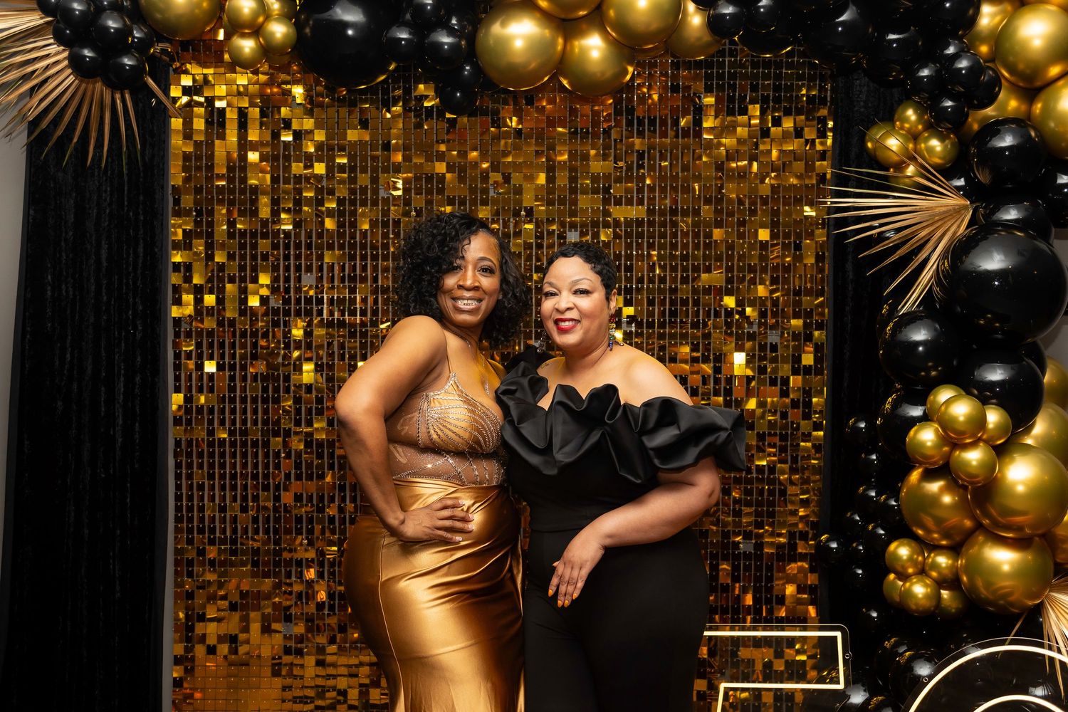 Two guests pose in front of a gold sequin backdrop with black and gold balloon decorations.
