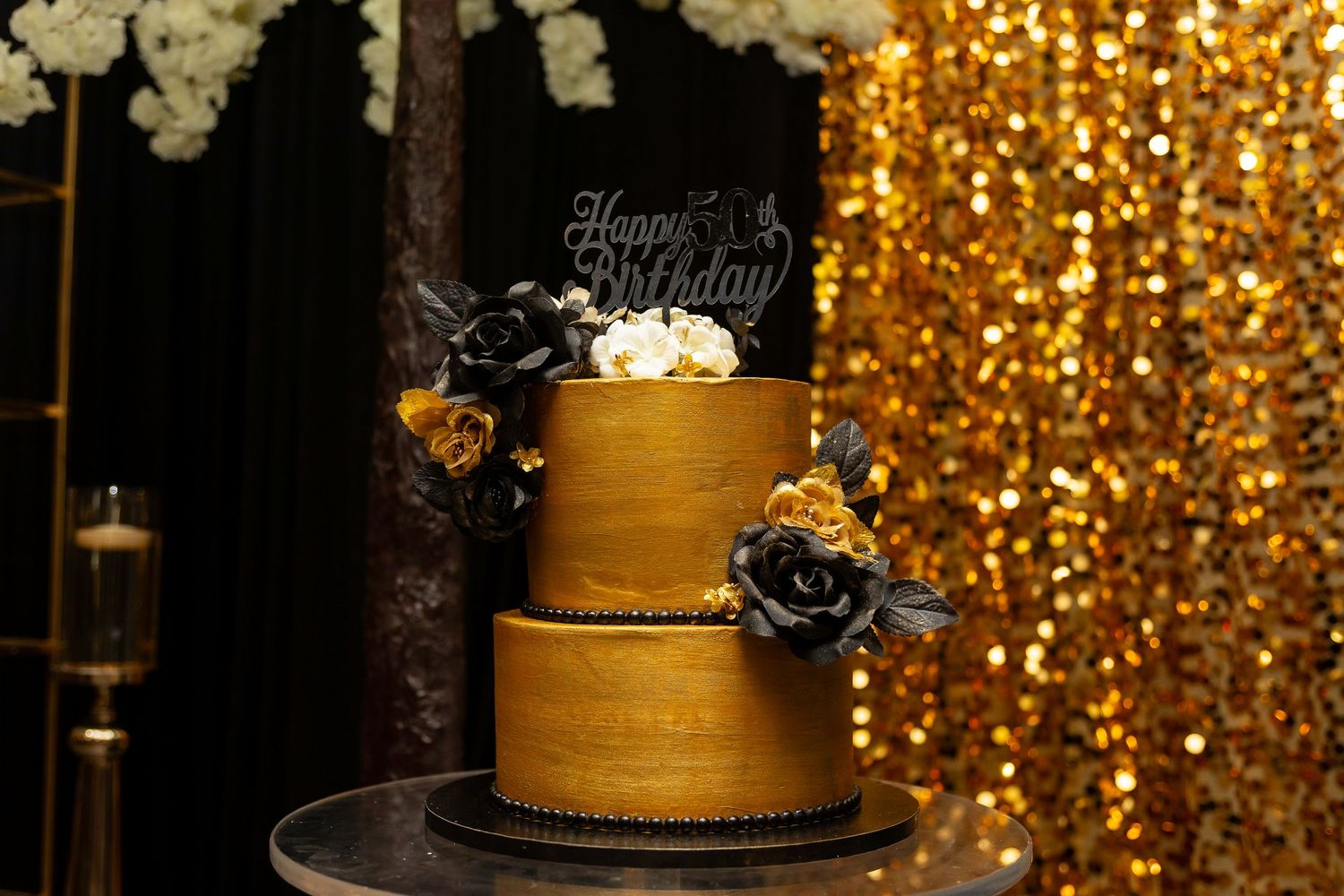 Elegant gold tiered birthday cake with black roses against a sparkly background.