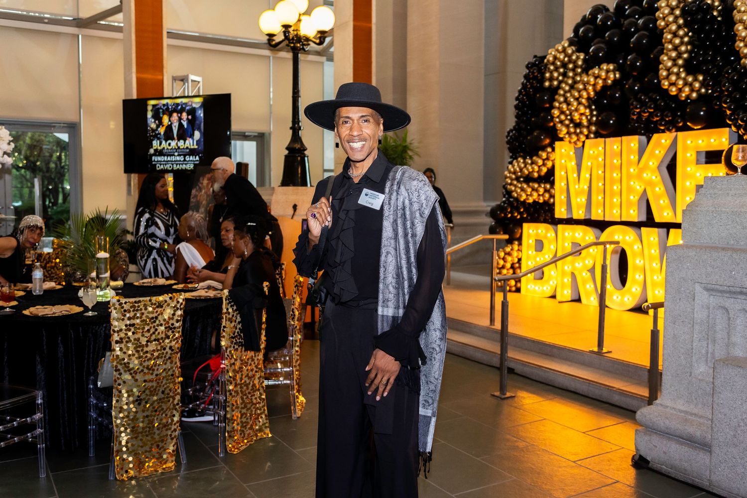 A guest in stylish attire poses near decorative gold signage at an evening social event.