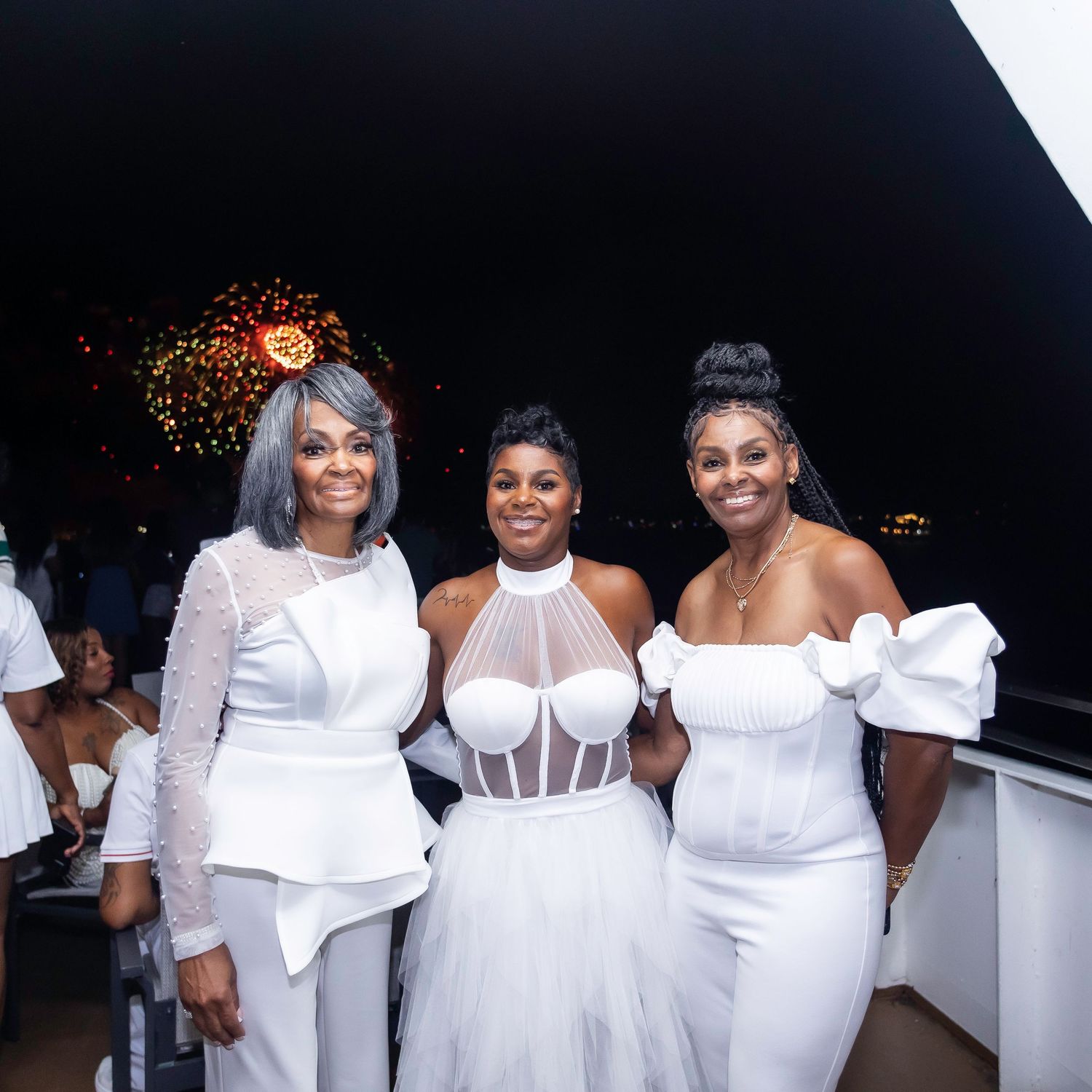Three women in elegant white dresses pose together at an all-white party celebration.