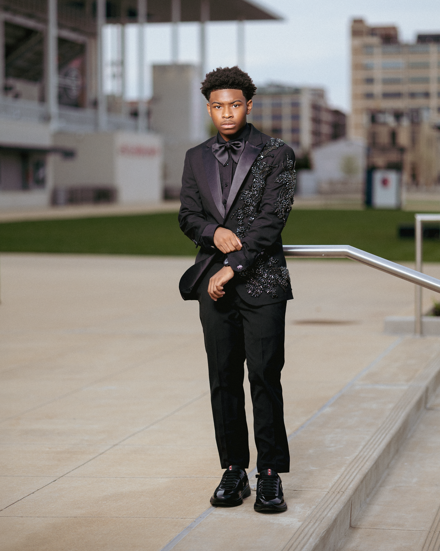 Young man in elegant black sequined tuxedo jacket with bow tie poses on steps in urban setting.