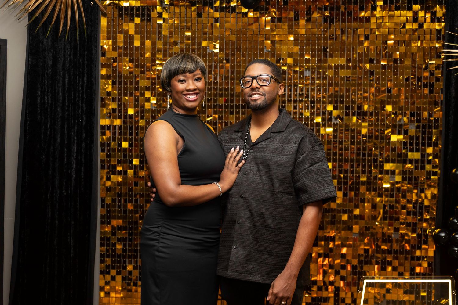 A couple poses together against a shimmering gold sequin backdrop at an event.
