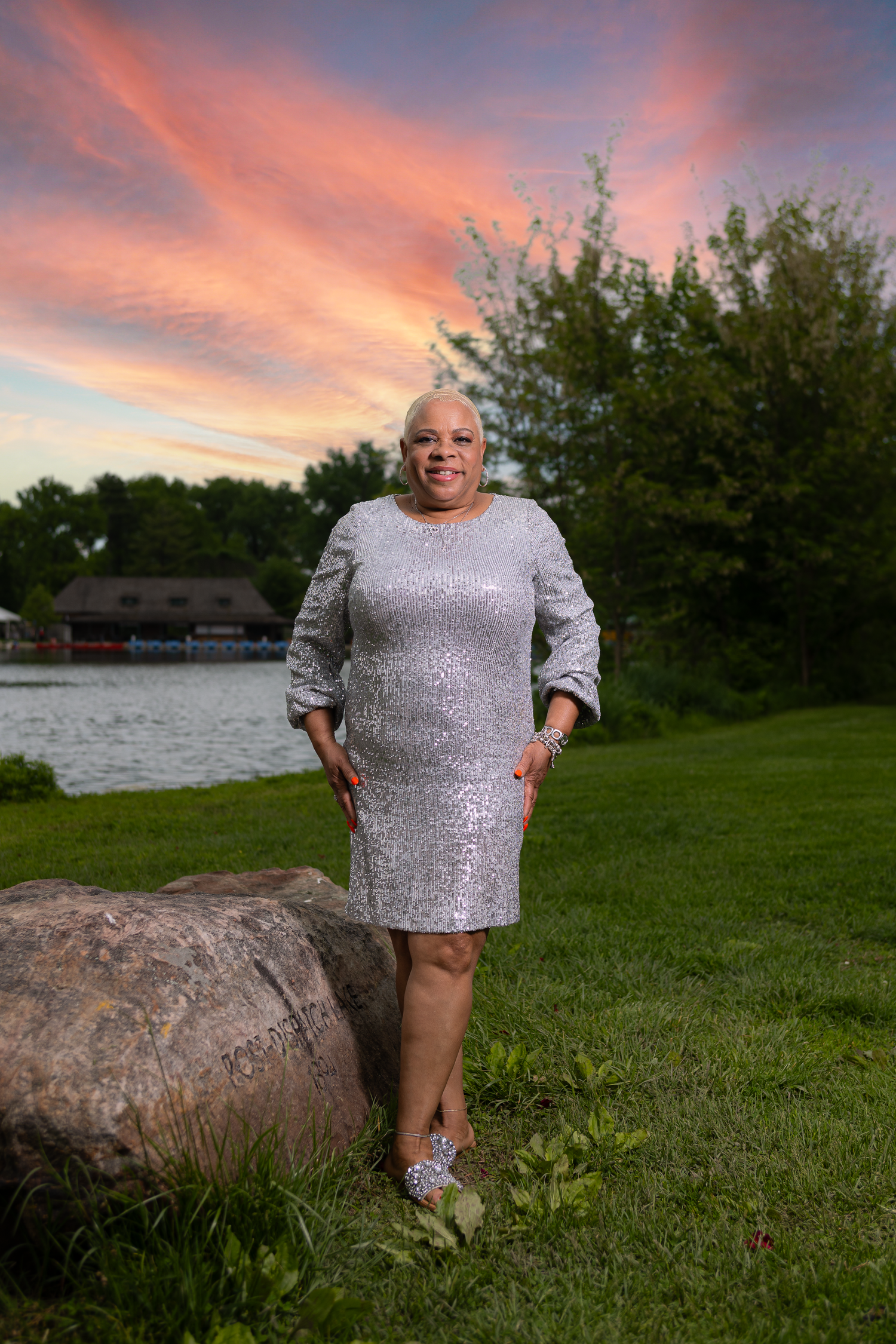 Person in glittery dress poses by lake during colorful sunset.