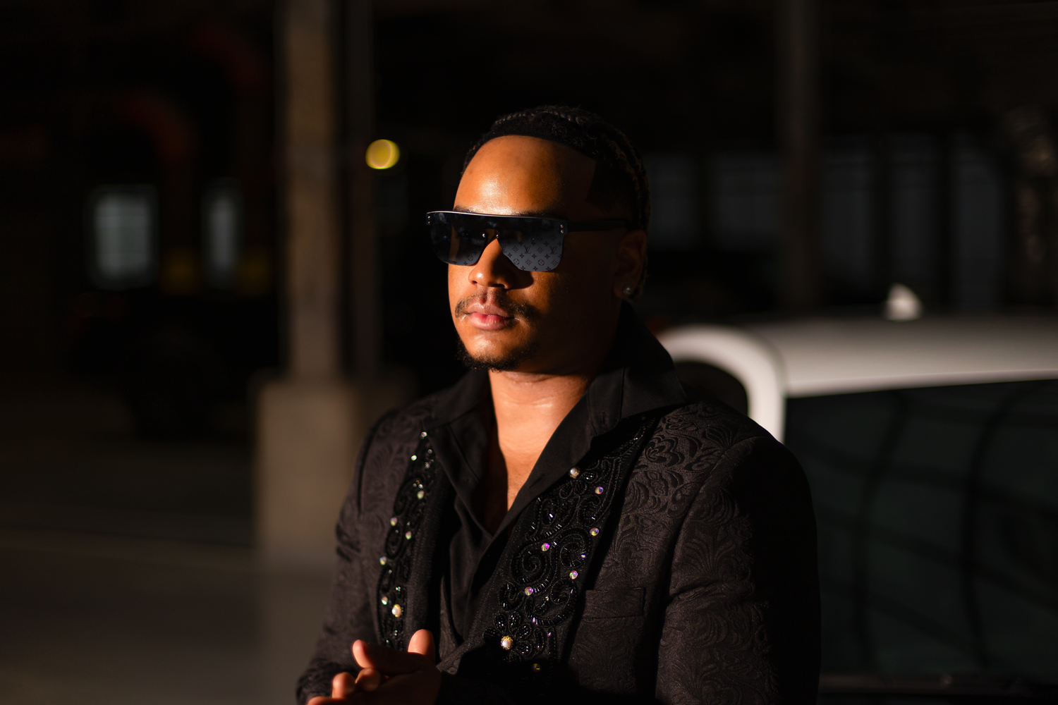 Fashion model in dark clothing strikes a pose next to luxury vehicle in moody lighting.