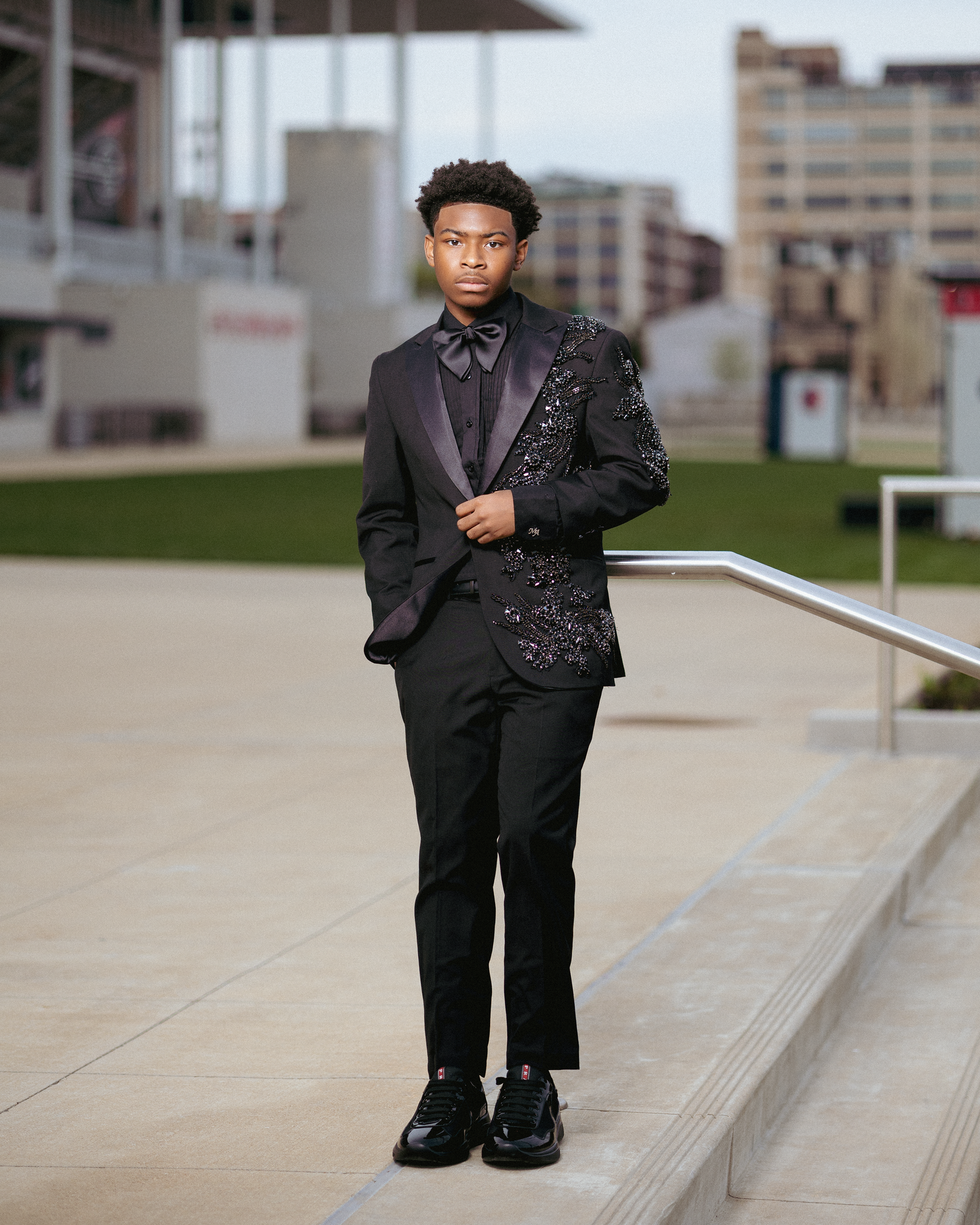 Young man in black sequined tuxedo jacket with bow tie poses on outdoor steps in urban setting.