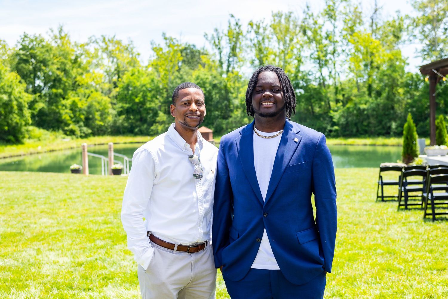 Two friends stand together outdoors near a lake with trees in background.