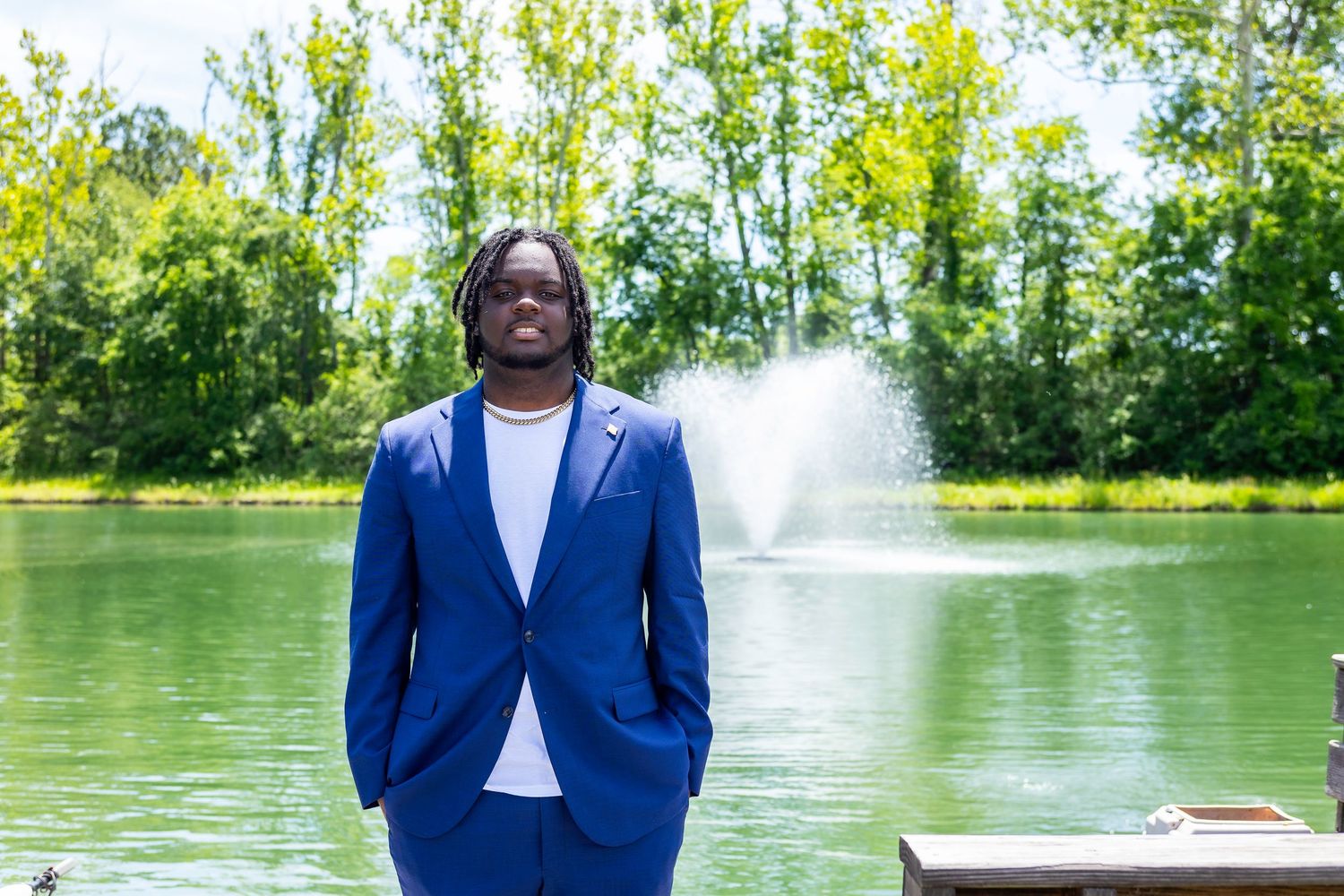 Person in blue suit stands by scenic lake with fountain spray in background.