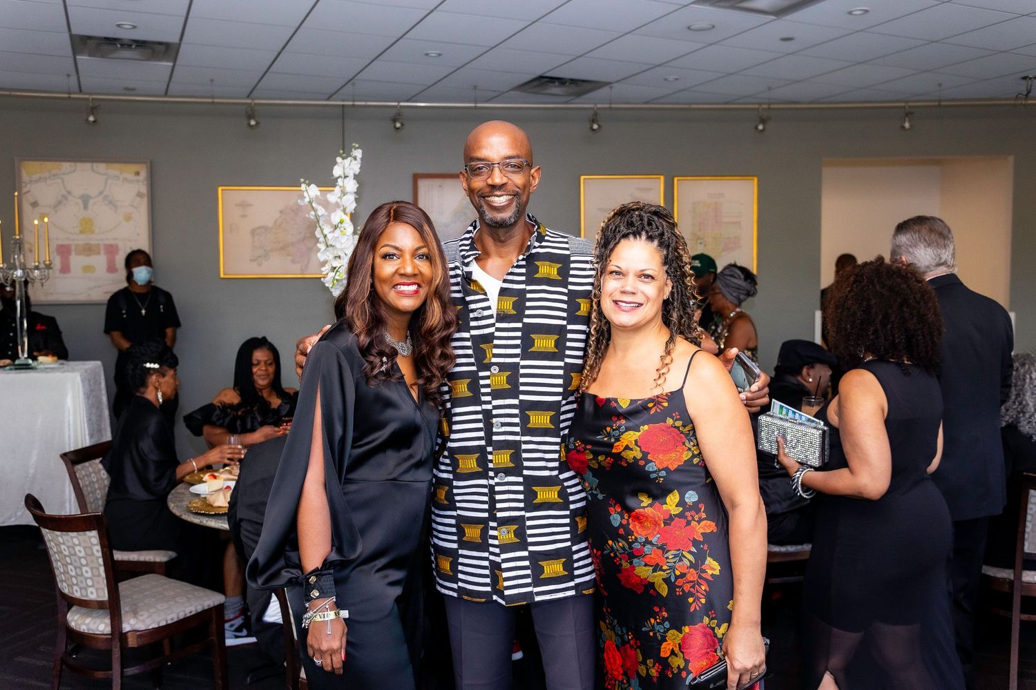 Three people pose together at a social event in a dining room with other guests visible in the background.