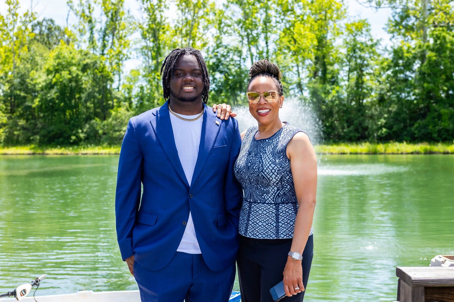 Two people pose together by a lake with a decorative fountain in the background.