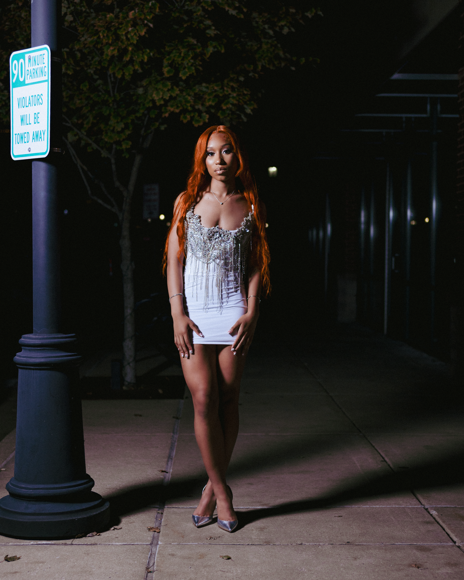 Woman in white beaded mini dress and silver heels with long red hair posing on urban sidewalk at night.