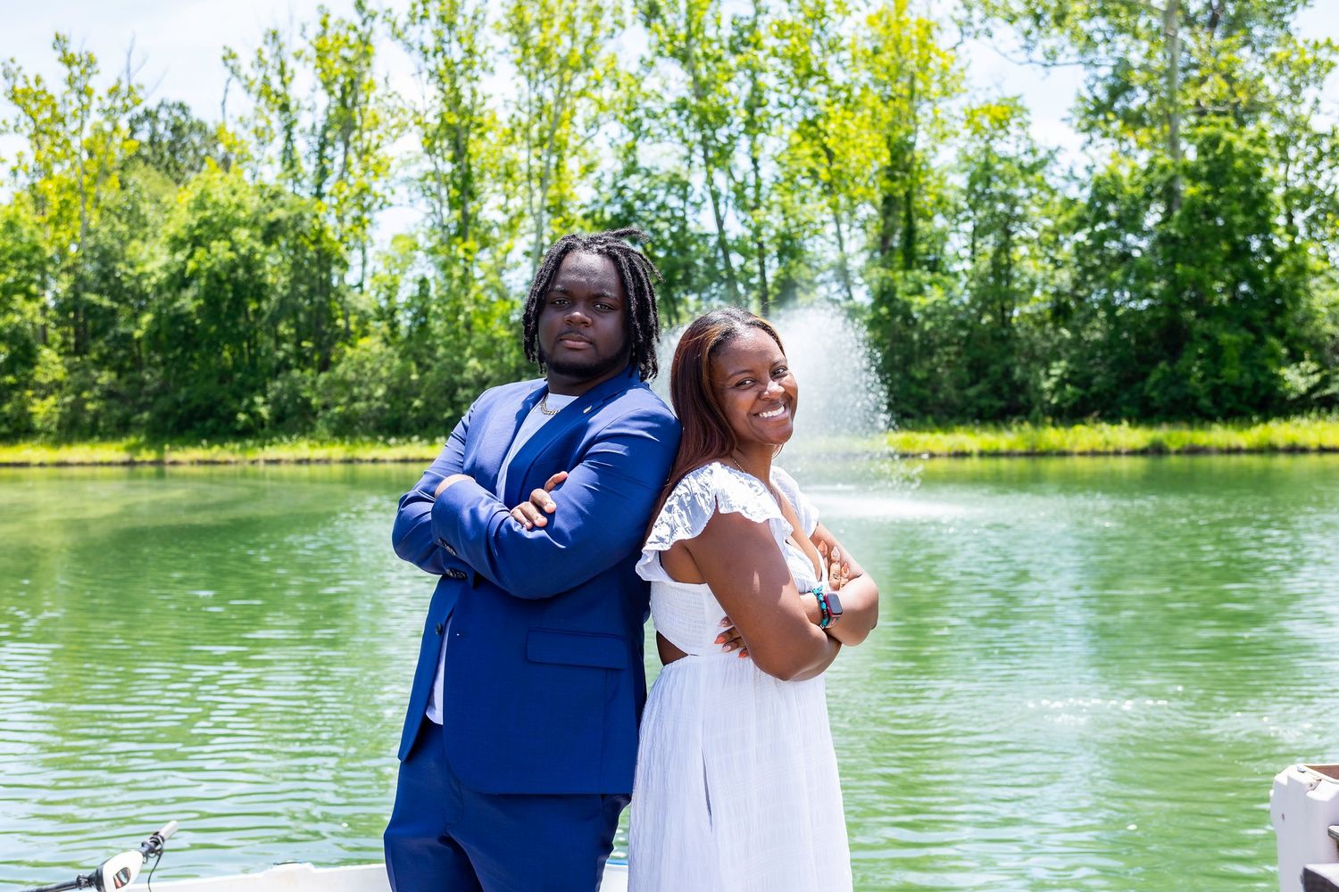 Two people stand back-to-back in formal attire by a lake with fountain.