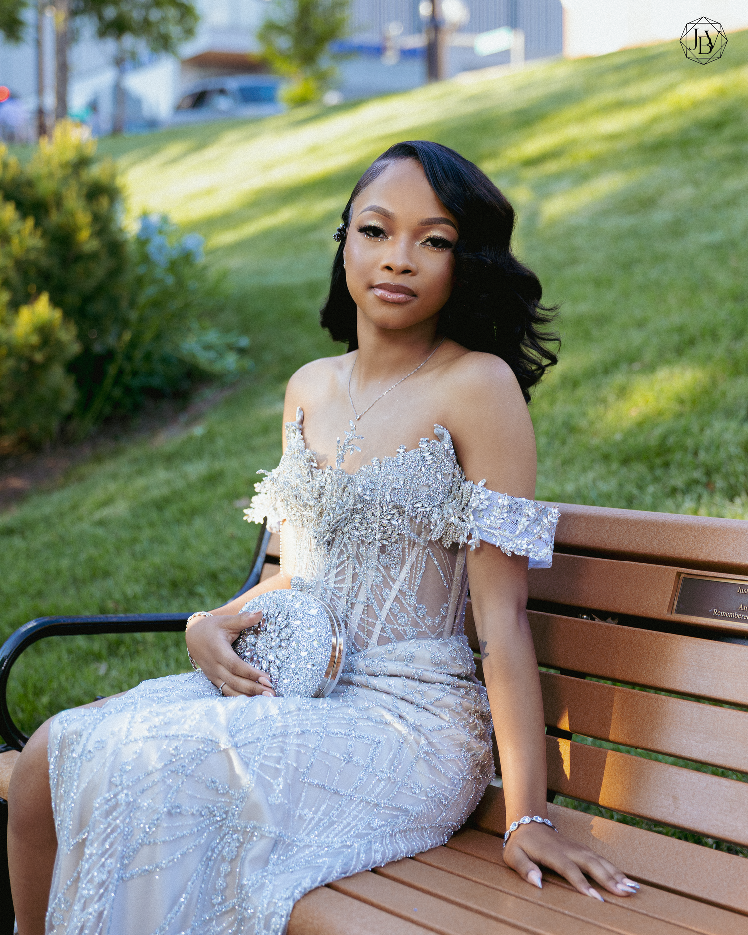 Young woman in elegant silver beaded off-shoulder gown sitting on a wooden bench outdoors.