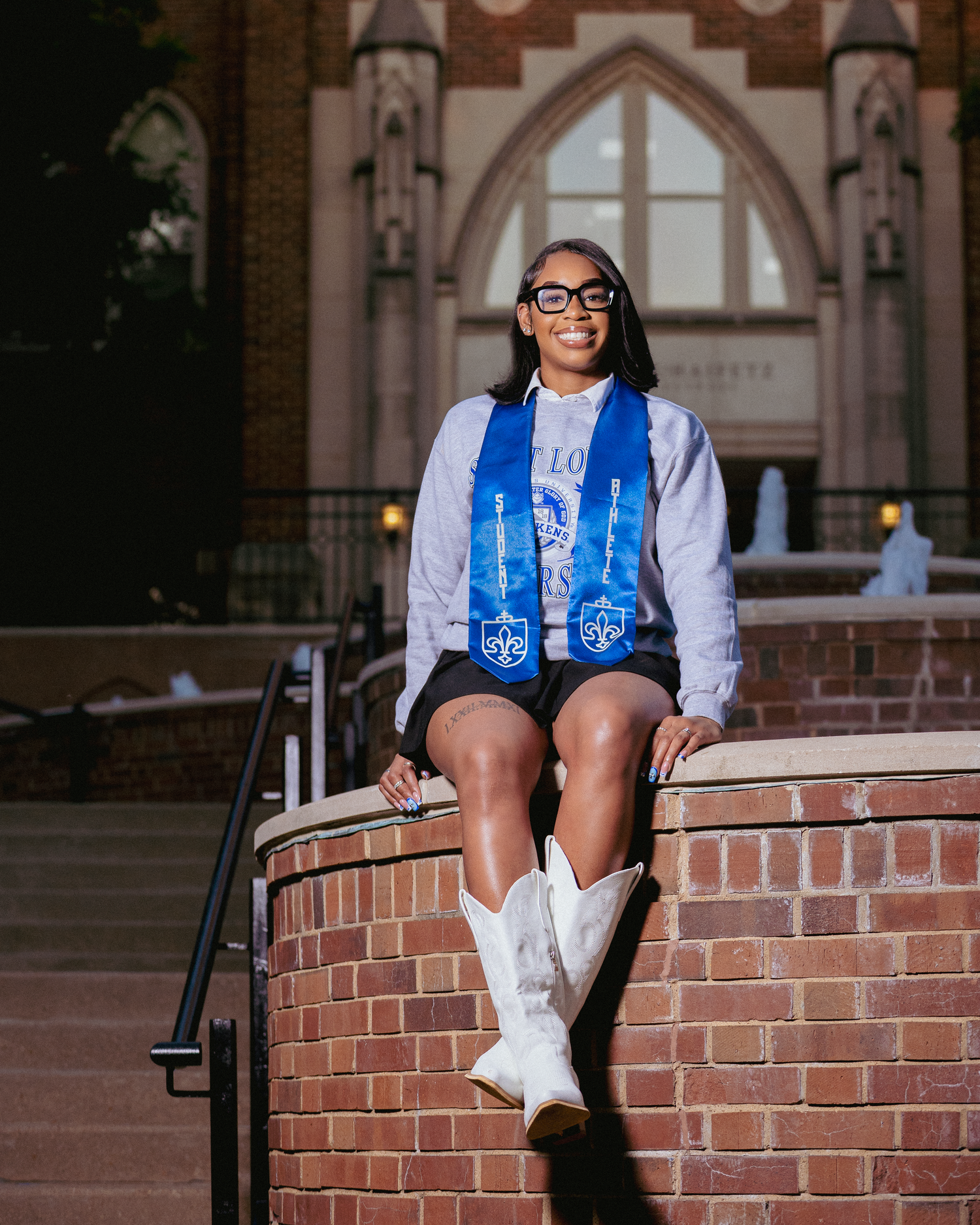 A smiling graduate sits on brick steps wearing a blue stole and white cowboy boots in front of a university building.