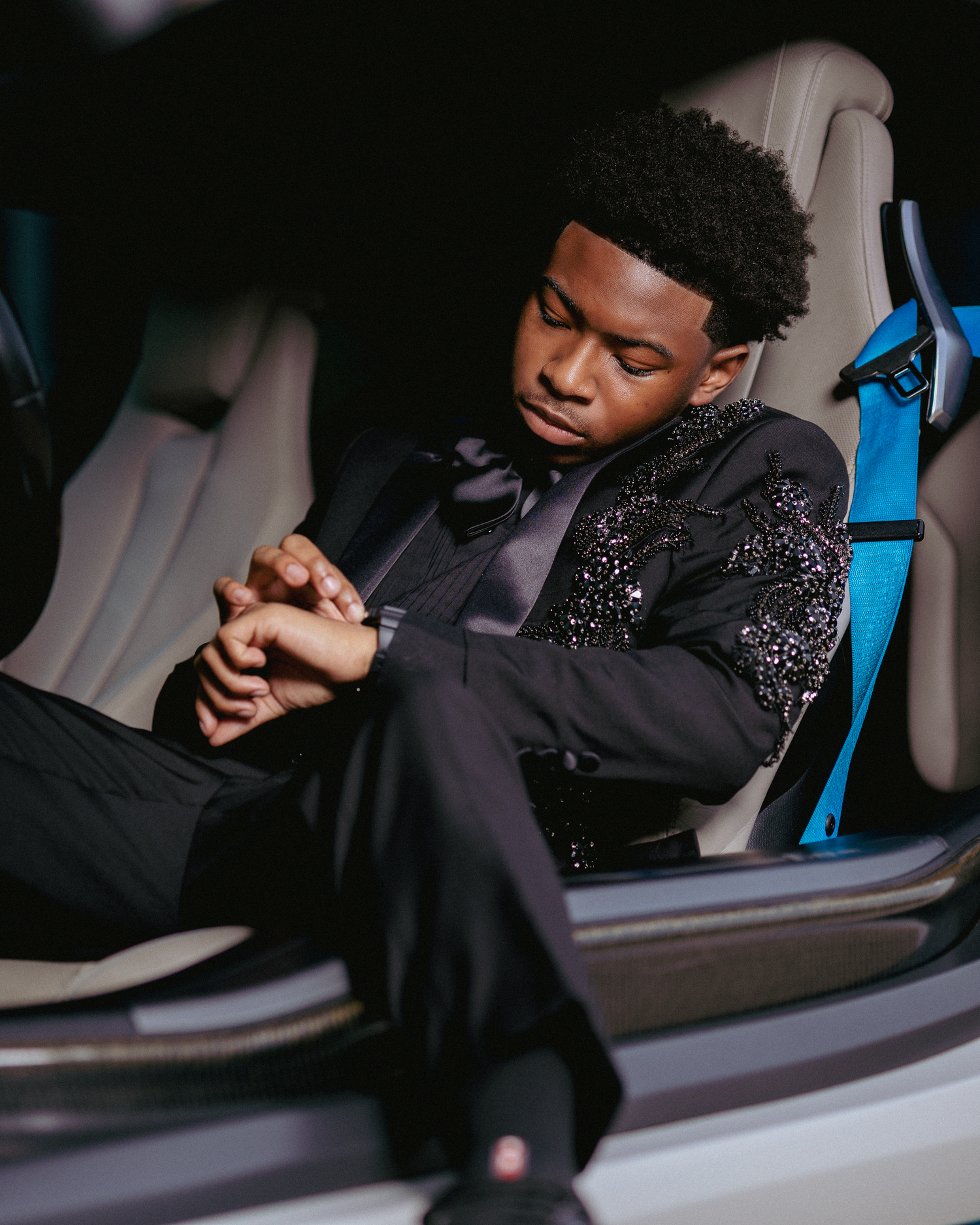Young man in a black sequined suit sitting in a luxury car with blue seatbelt, looking at his watch.