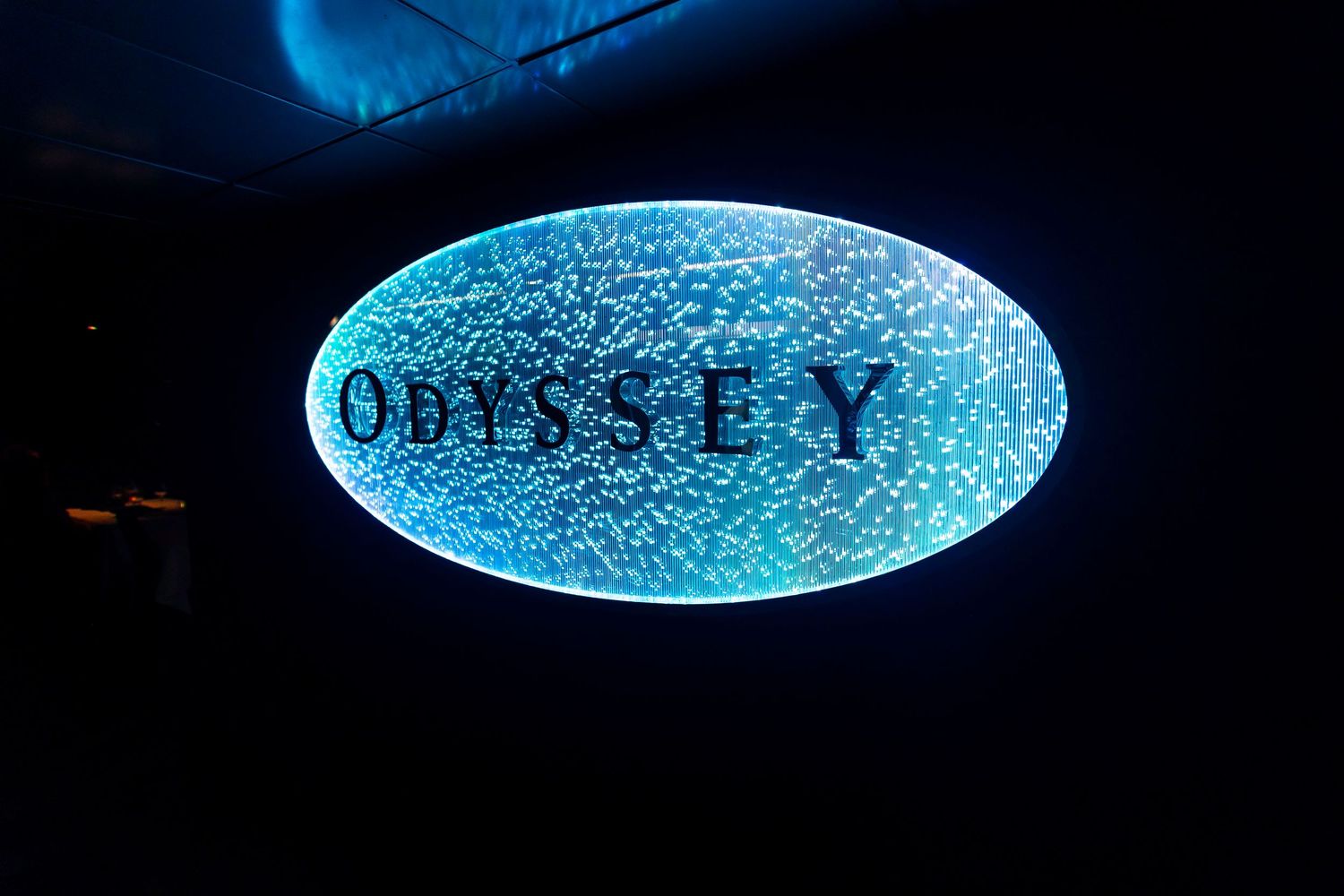 Illuminated blue oval sign reading 'Odyssey' with crackled texture effect against dark background.