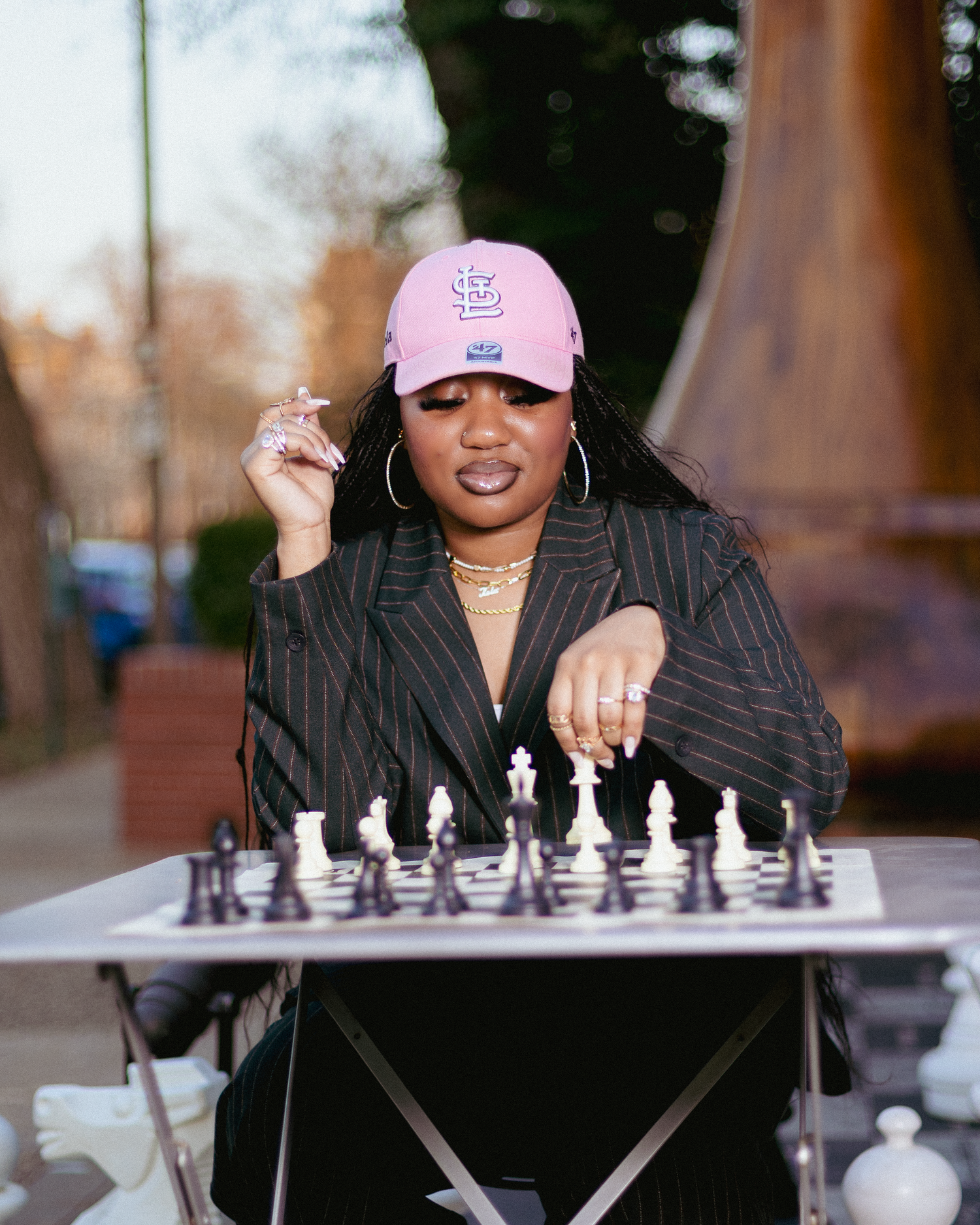 Woman in pink cap and black blazer playing chess outdoors at a table.