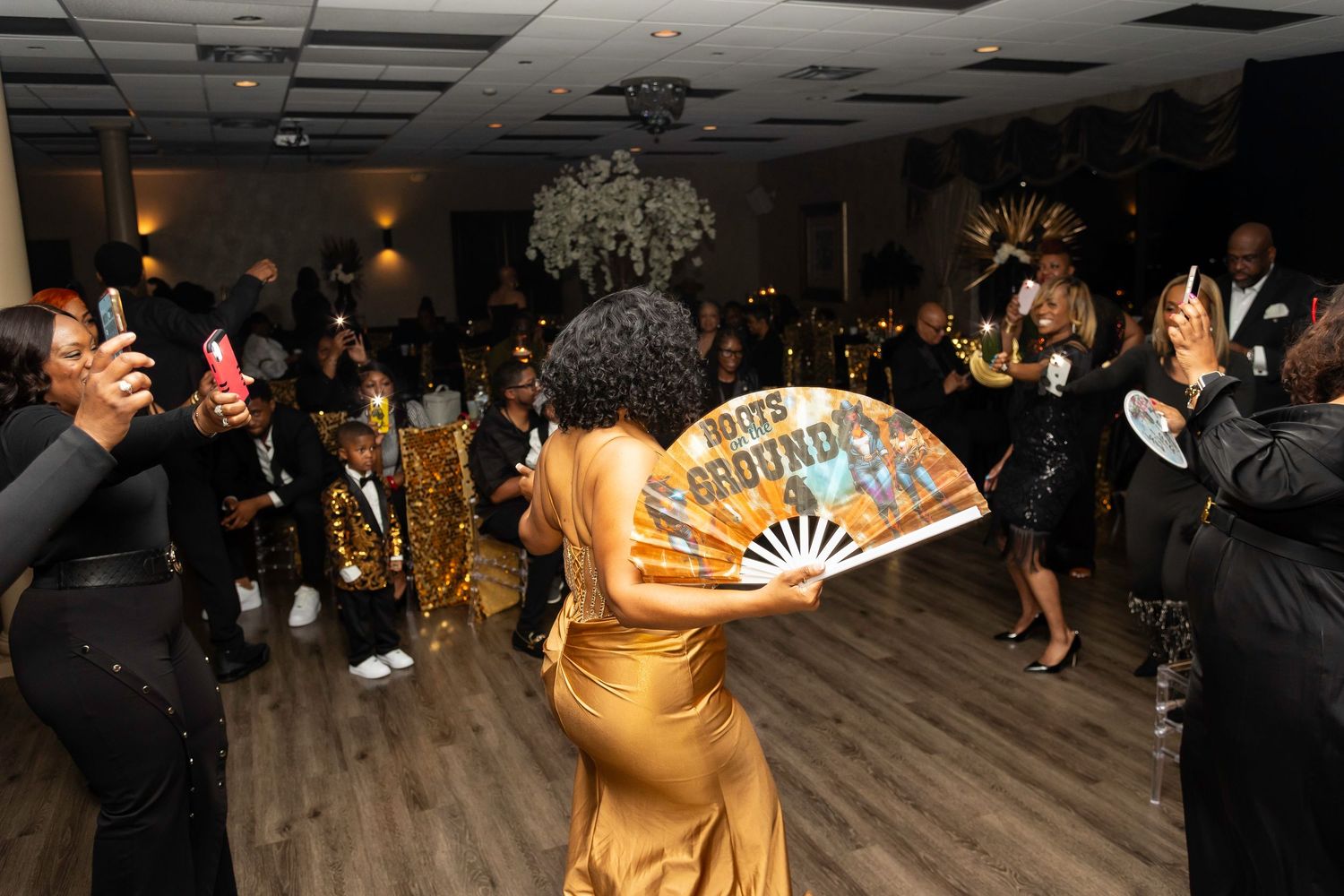 Guest in gold dress performs with a fan on the dance floor as others watch and take photos.
