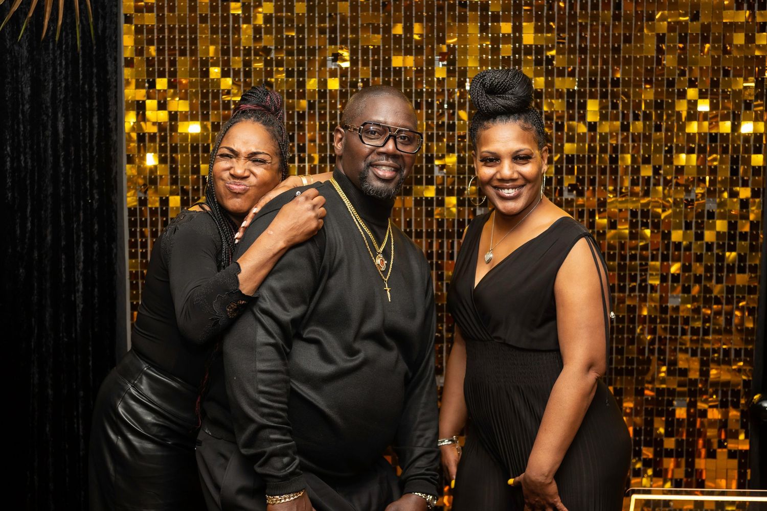 Party guests pose against a glamorous gold sequin wall with black and gold balloons.