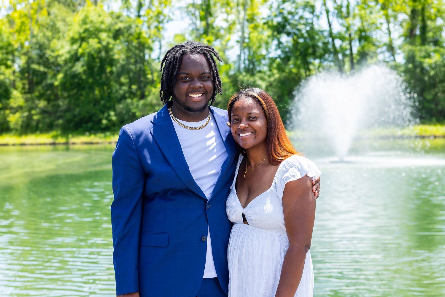 Couple poses together by scenic lake with water fountain in the background.