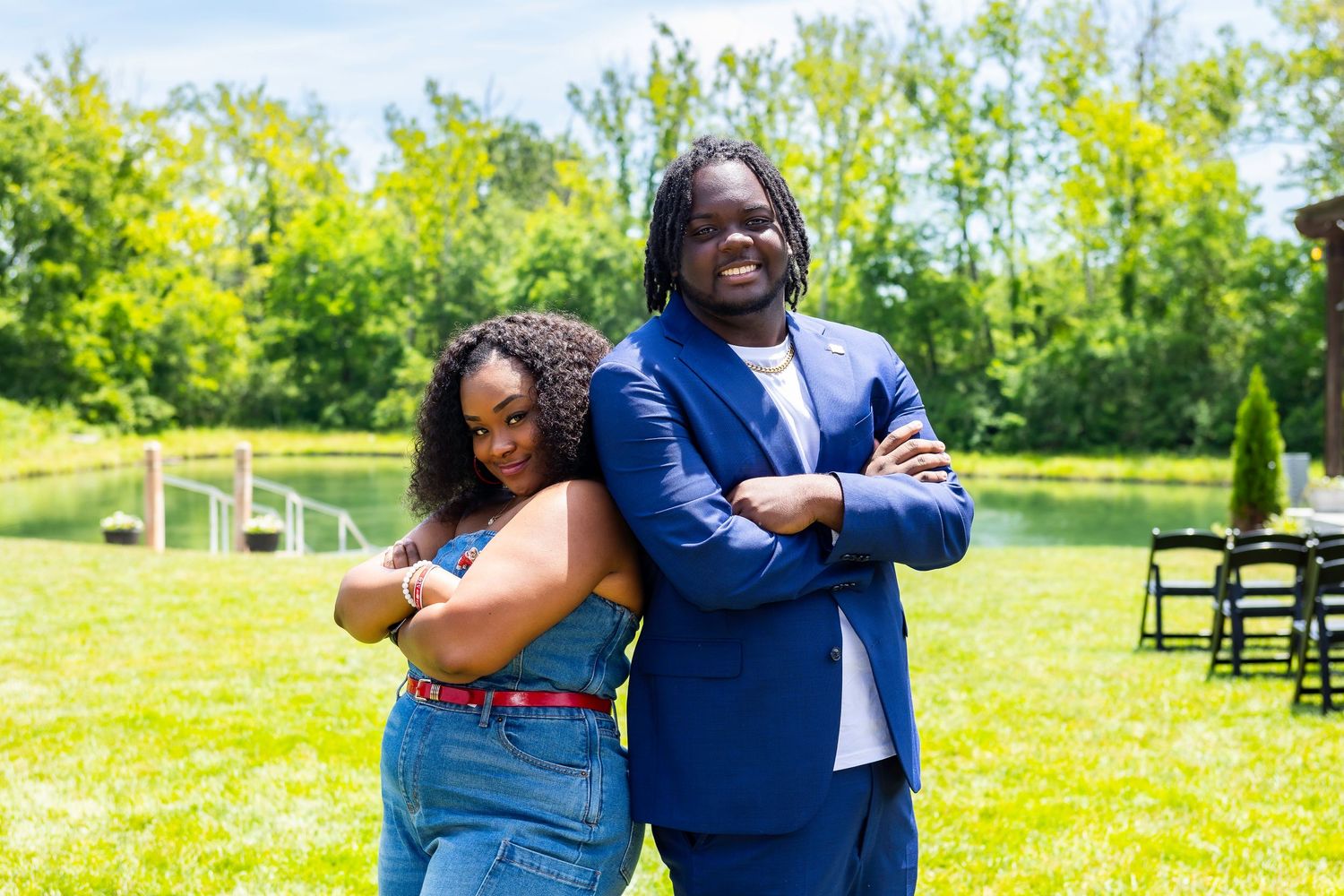 Two people pose together playfully in front of trees on a grassy lawn.