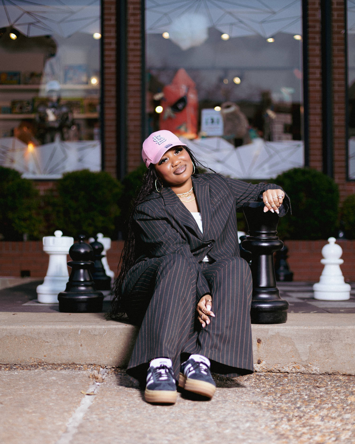 A stylish woman in a pinstripe suit and pink cap sits confidently among giant chess pieces outside a shop.