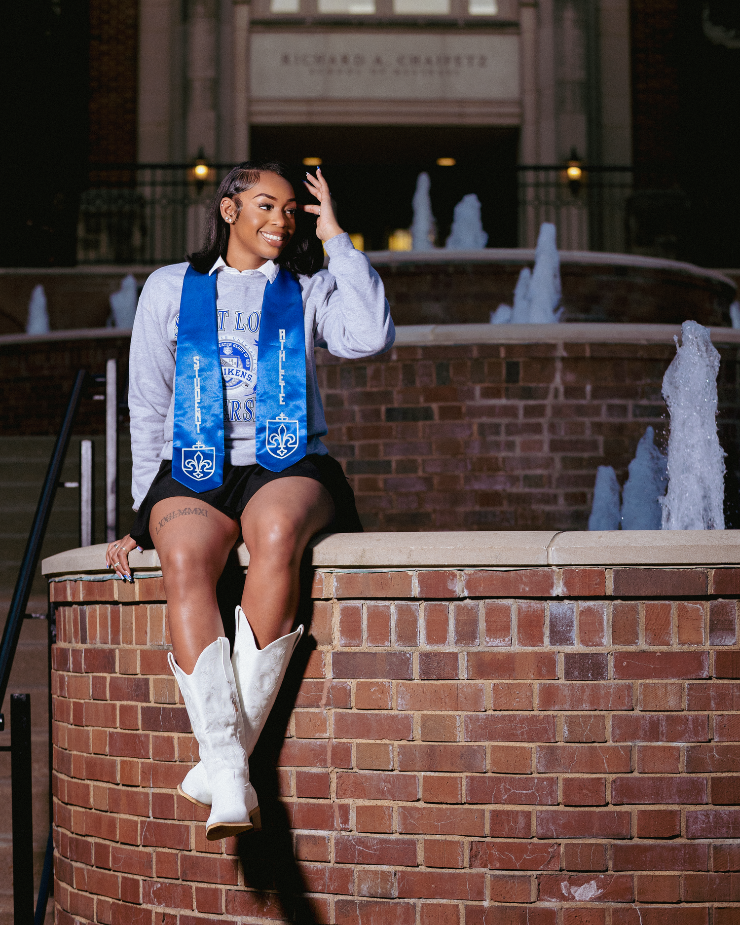 Graduate in blue stole and white cowboy boots poses on brick wall near fountain at university campus.
