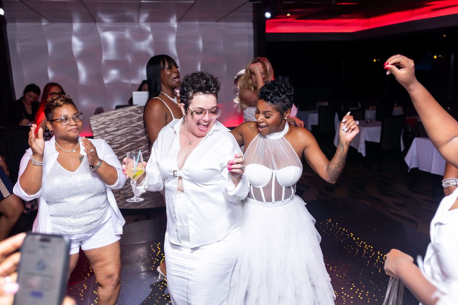 Party guests in white attire dancing and celebrating on a dance floor with festive lighting.