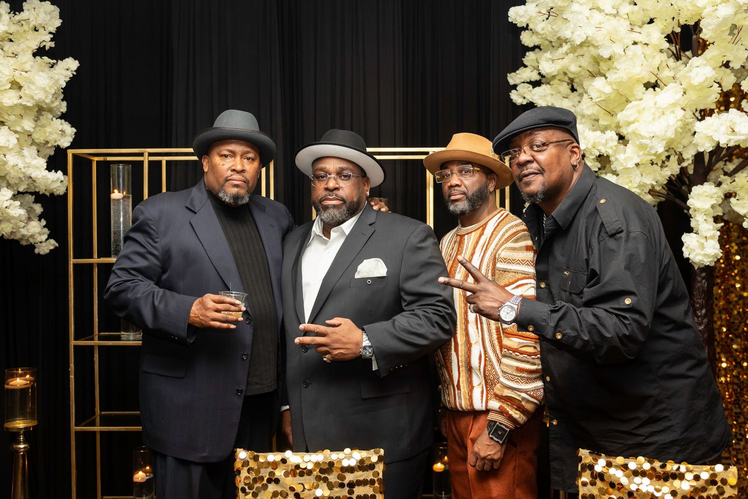 Four stylishly dressed guests pose together at an elegant event with gold decor and floral arrangements in the background.