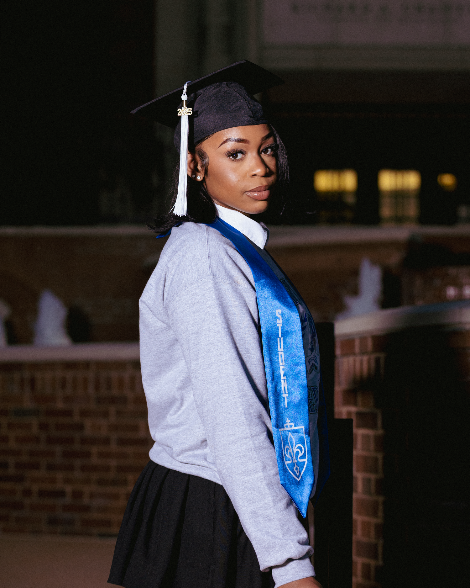 Young woman in graduation cap and blue stole poses proudly outdoors at university campus ceremony.