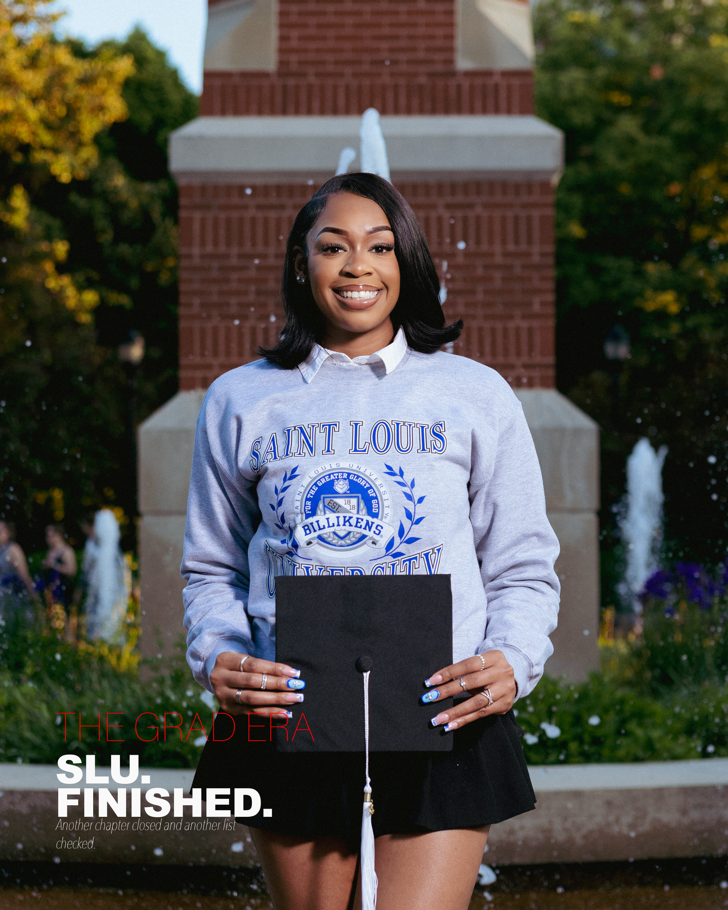 Saint Louis University graduate smiles proudly in SLU sweatshirt, holding diploma at campus fountain.