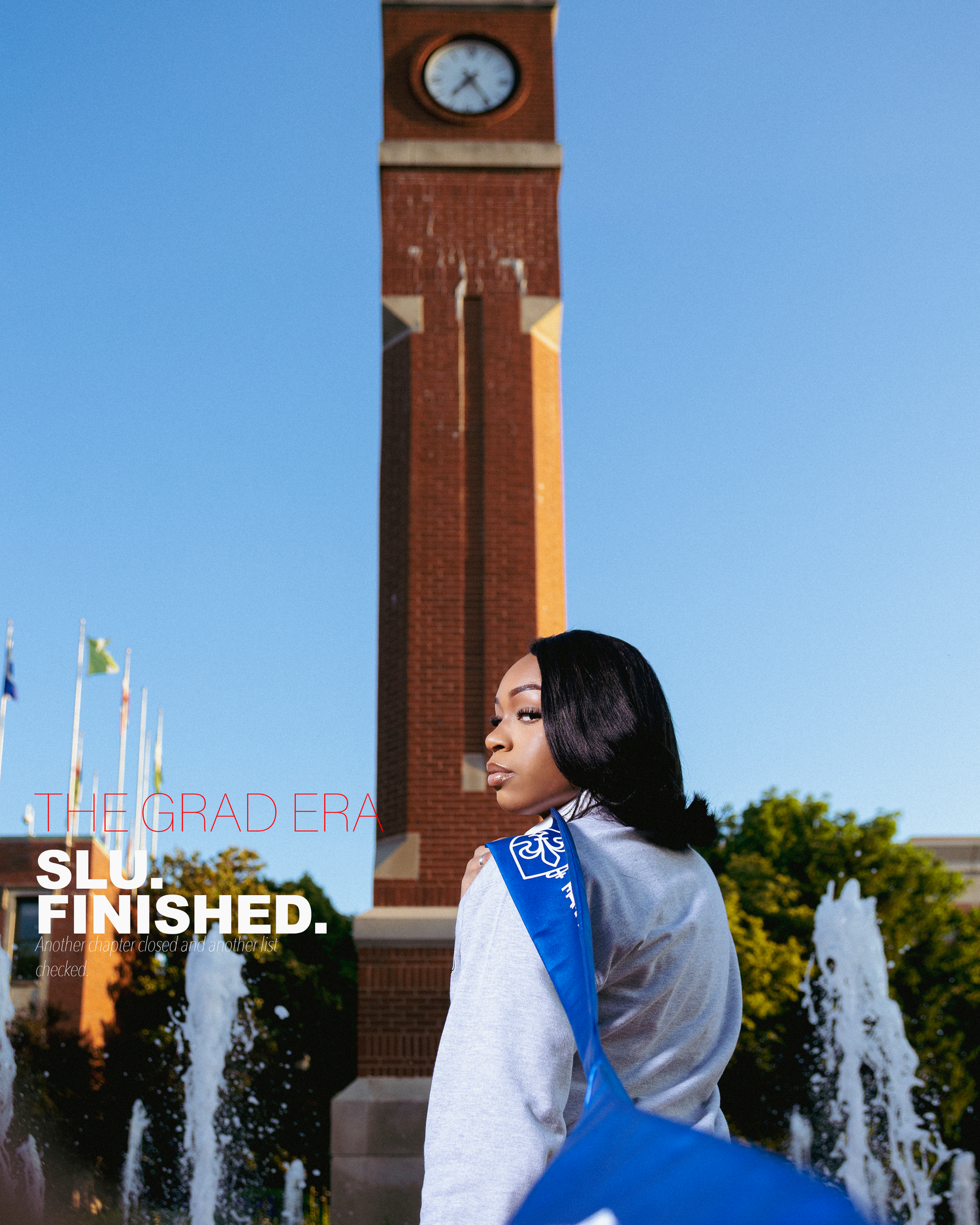 A graduate poses in a blue sash near a brick clock tower with fountains, celebrating her achievement.