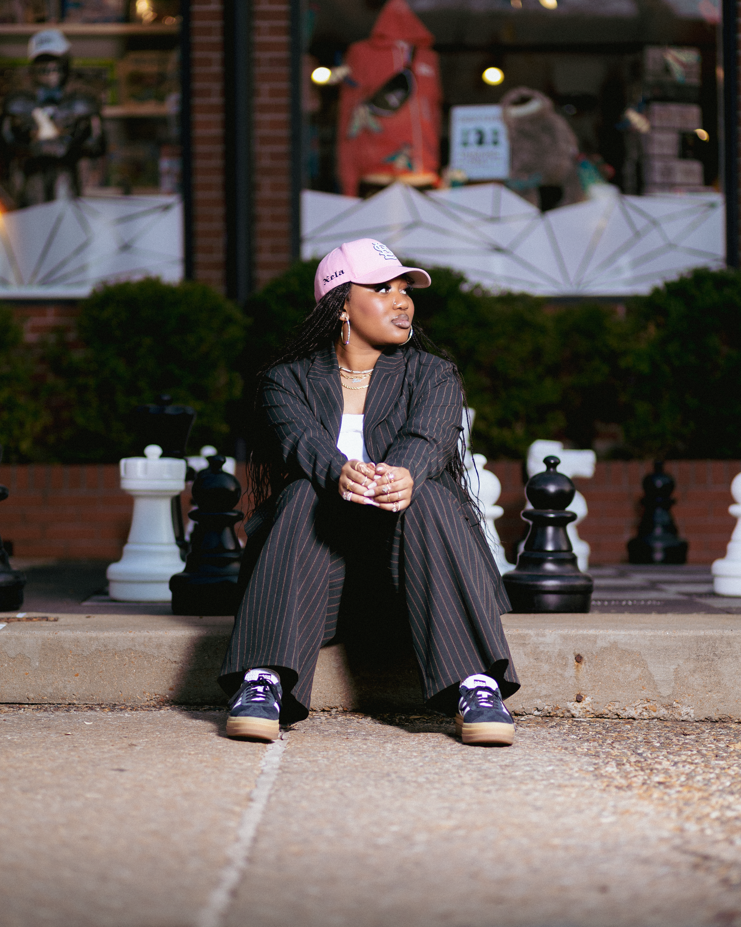 Person in pinstripe suit and pink cap sits on pavement near oversized chess pieces outside a brick building.