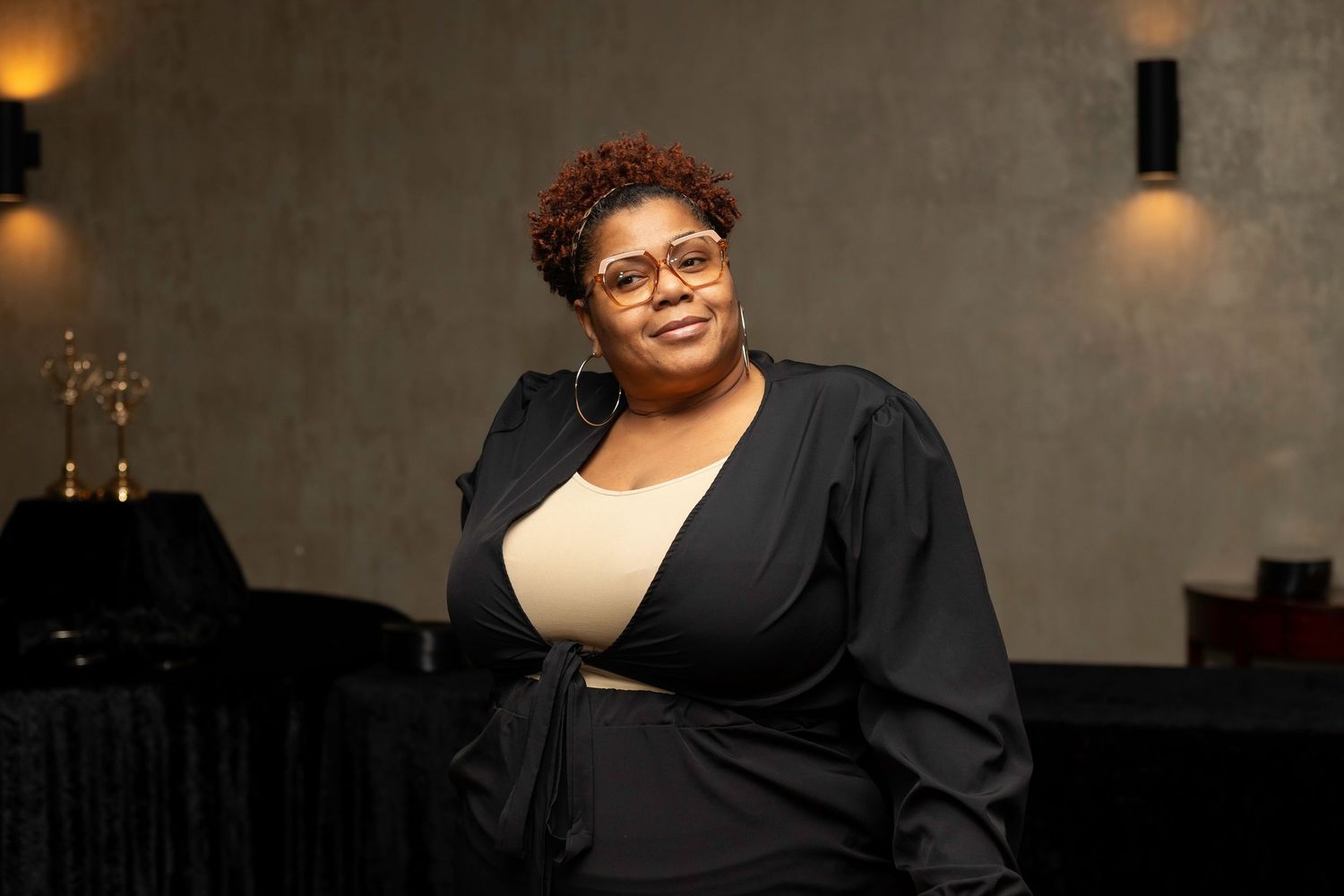 Person in formal black attire poses against neutral wall with warm lighting.
