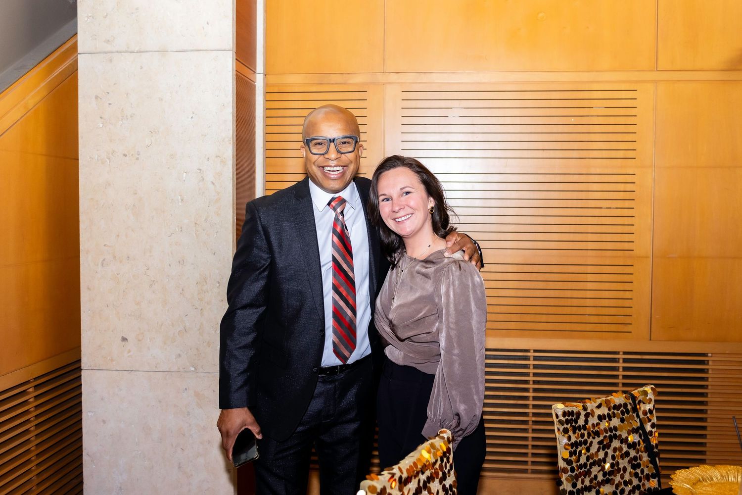 Two guests pose together in formal attire against a beige wall at an indoor event.