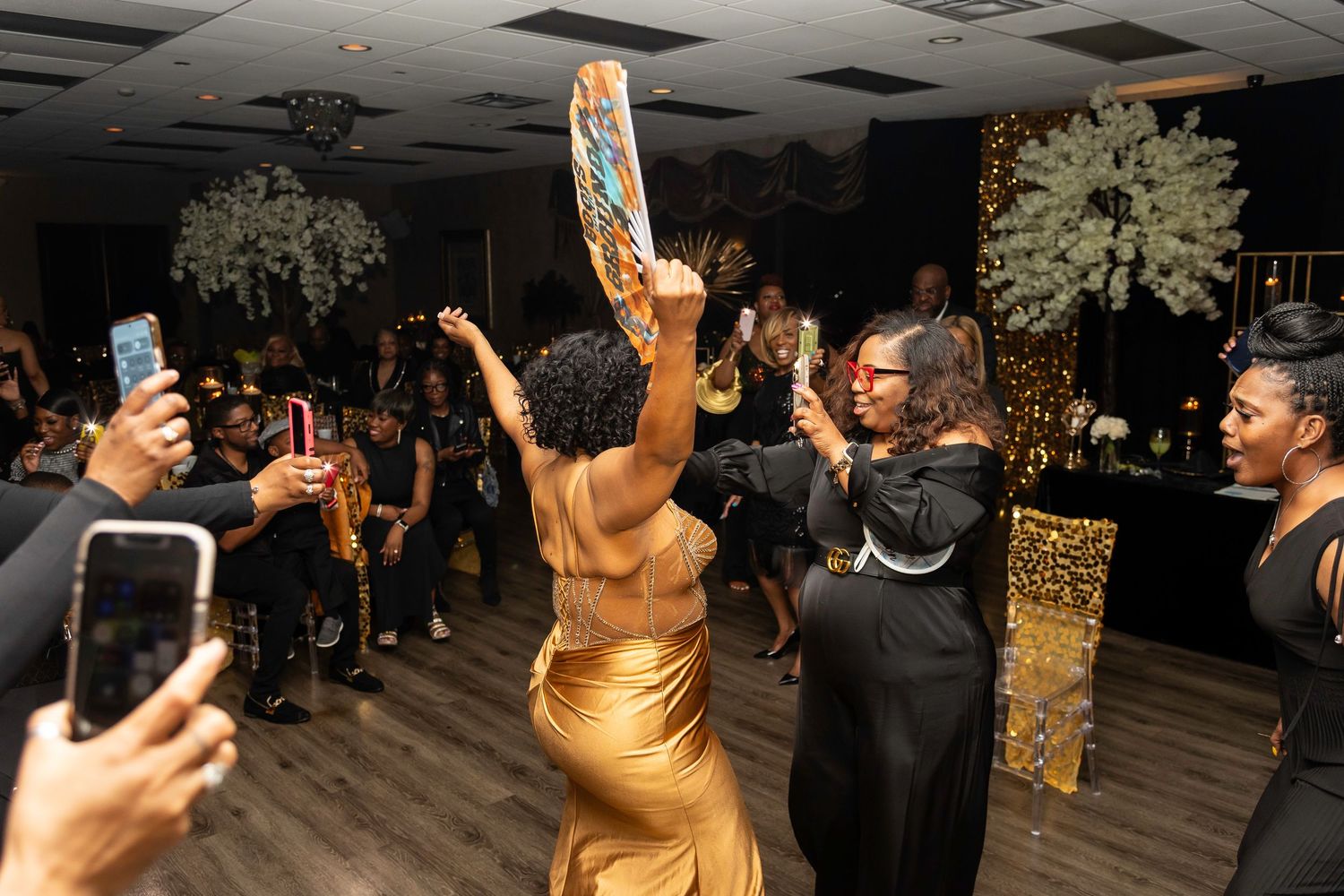 Guest in gold dress dancing while others capture the moment on phones.