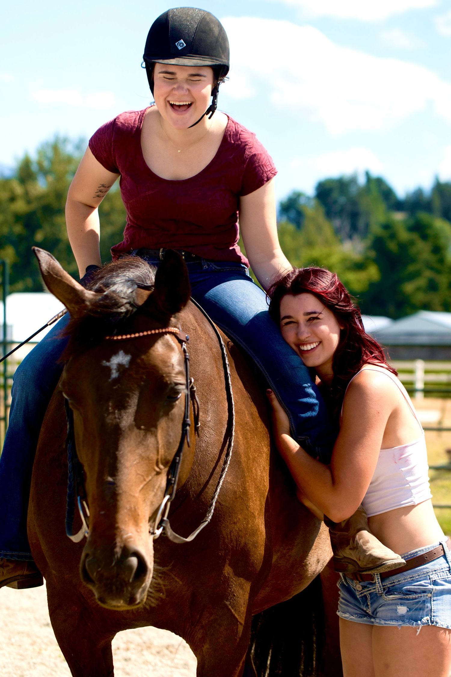 female in red shirt sitting on horse laughing and female on ground hugging her leg