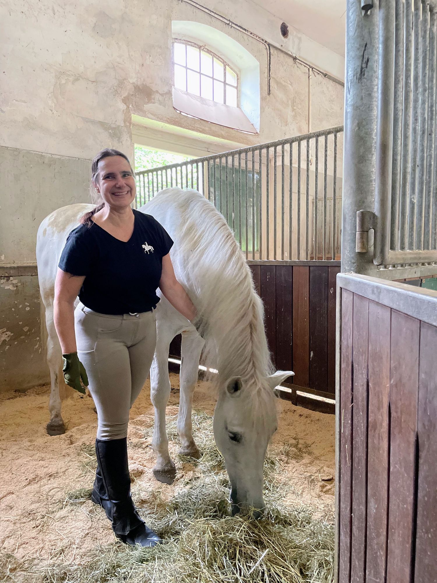 Laughing woman in a German box stall with grey horse that is eating hay, Karlsruhe, Egon von Neindorff Reitinstitut, equine