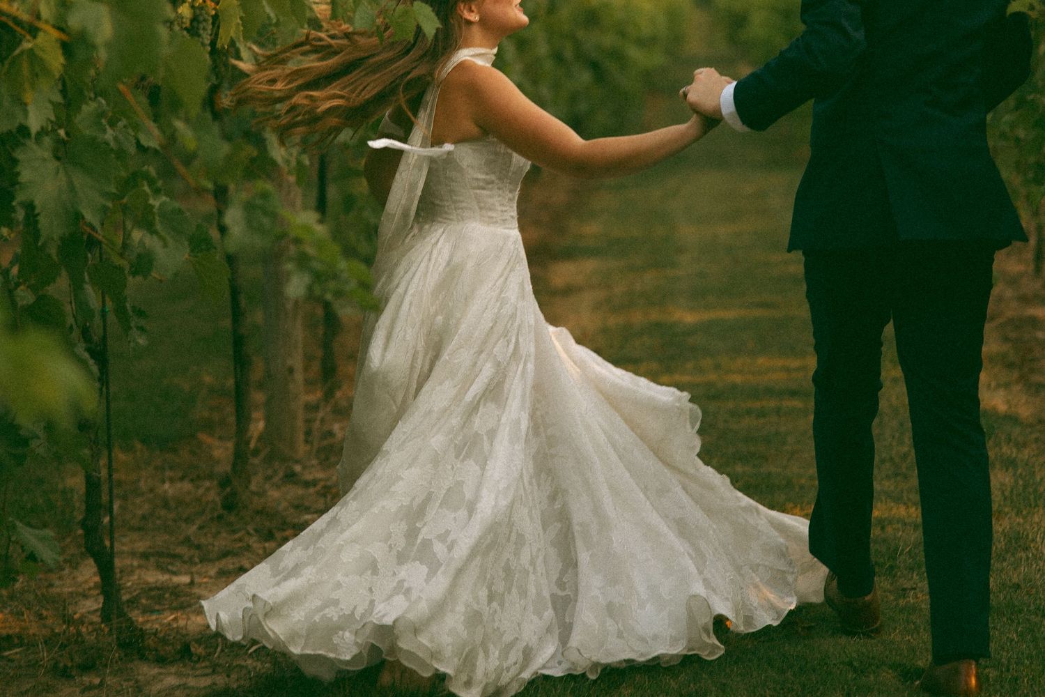 Bride in a flowing lace wedding gown twirls while holding the groom's hand among vineyard rows.