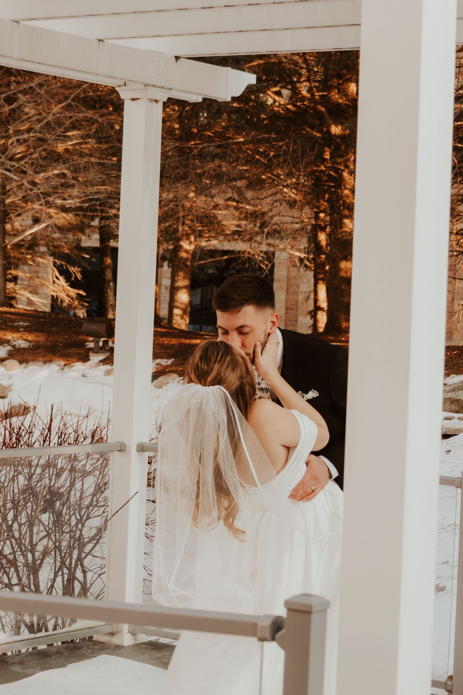 Groom in a black suit leans in to kiss his bride in a white gown and veil beside a white pergola in winter.
