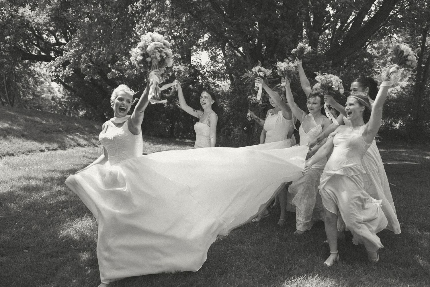 Black and white photo of bridesmaids cheering and raising bouquets as the bride's long gown flows across the grass.