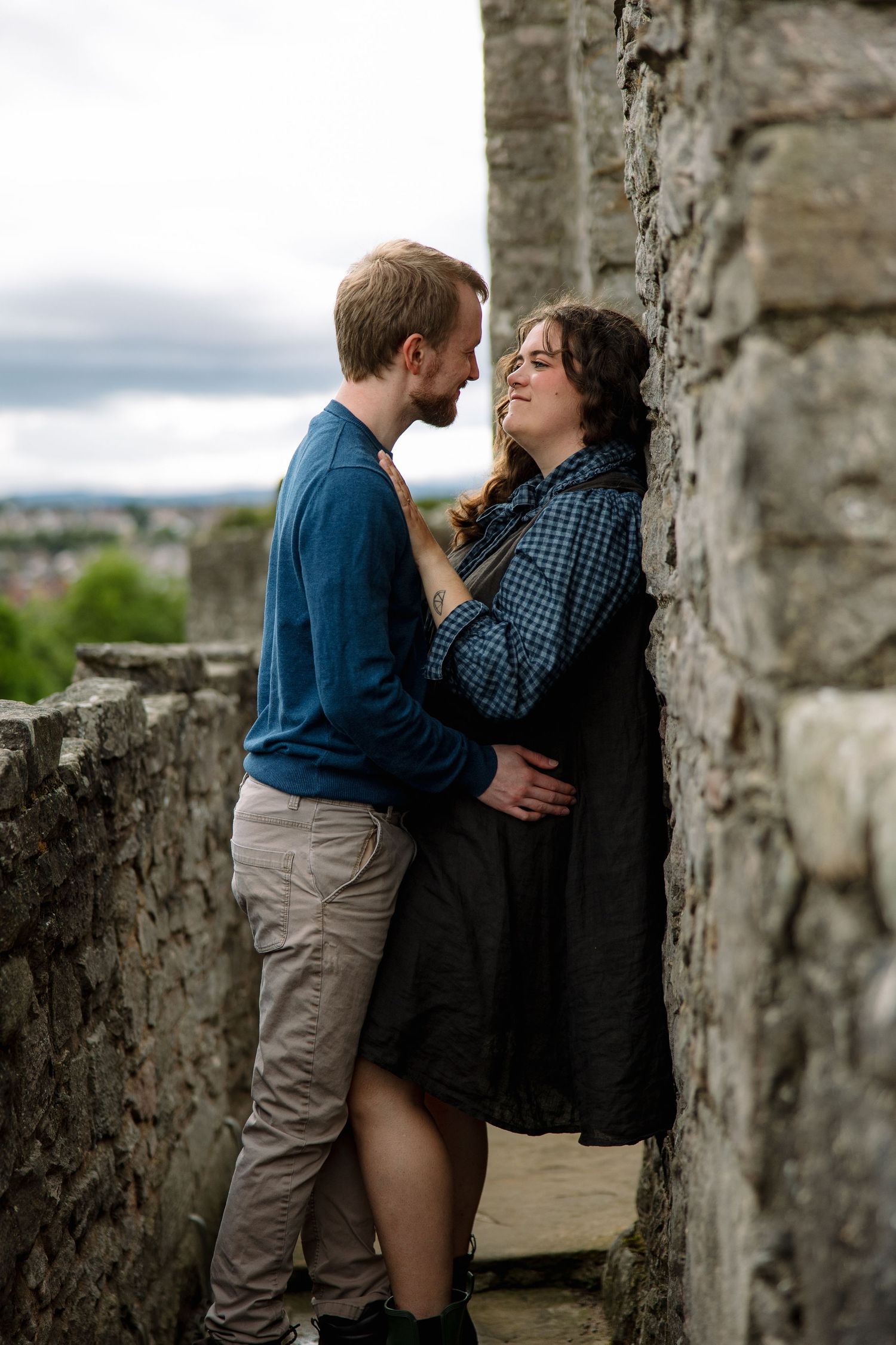 A couple embraces and smiles between stone castle walls, with a hazy green landscape and cloudy sky behind them.