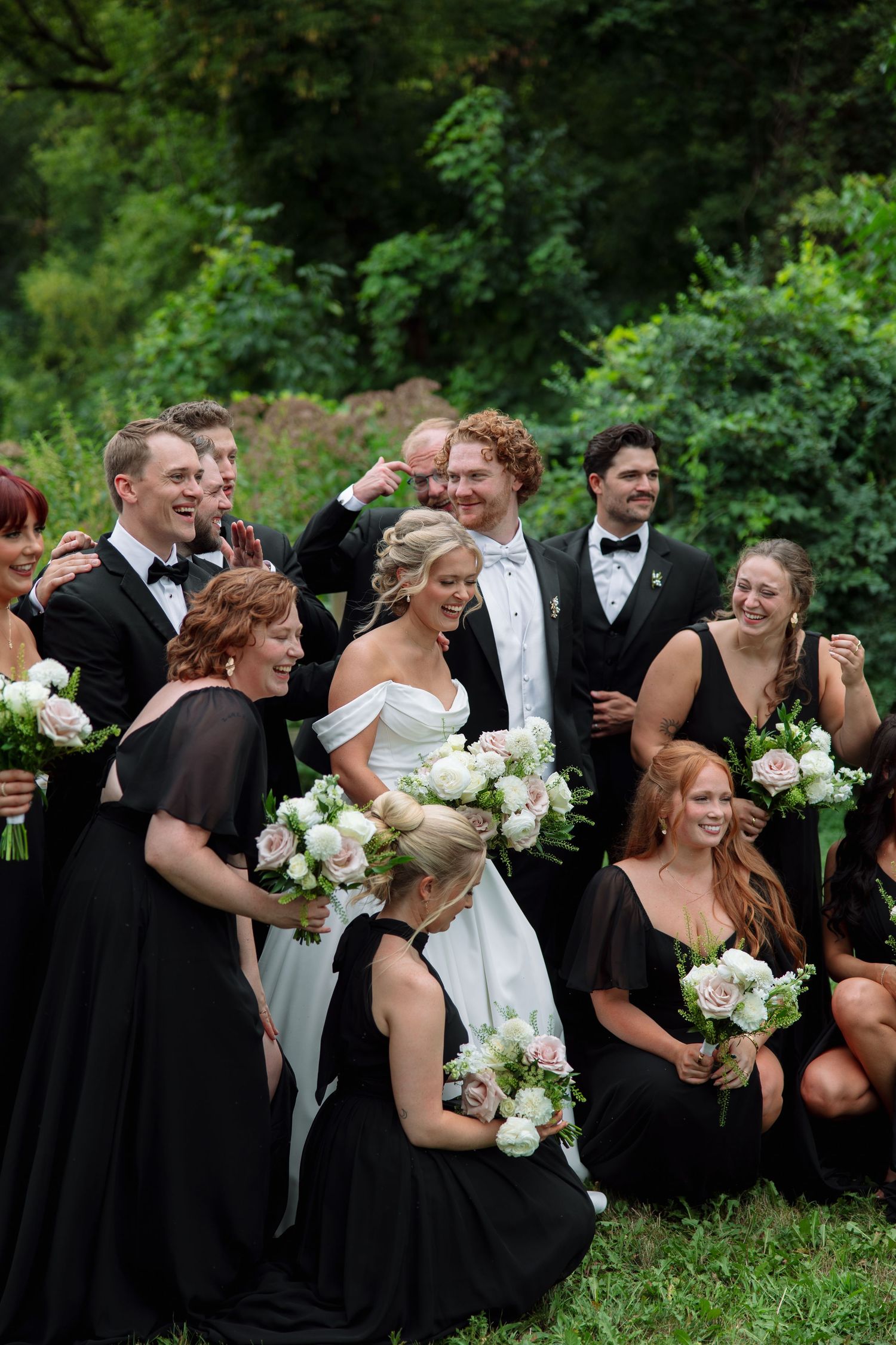 Bride in a white gown laughs with groomsmen in black tuxedos and bridesmaids in black dresses holding white and blush bouquets outdoors.